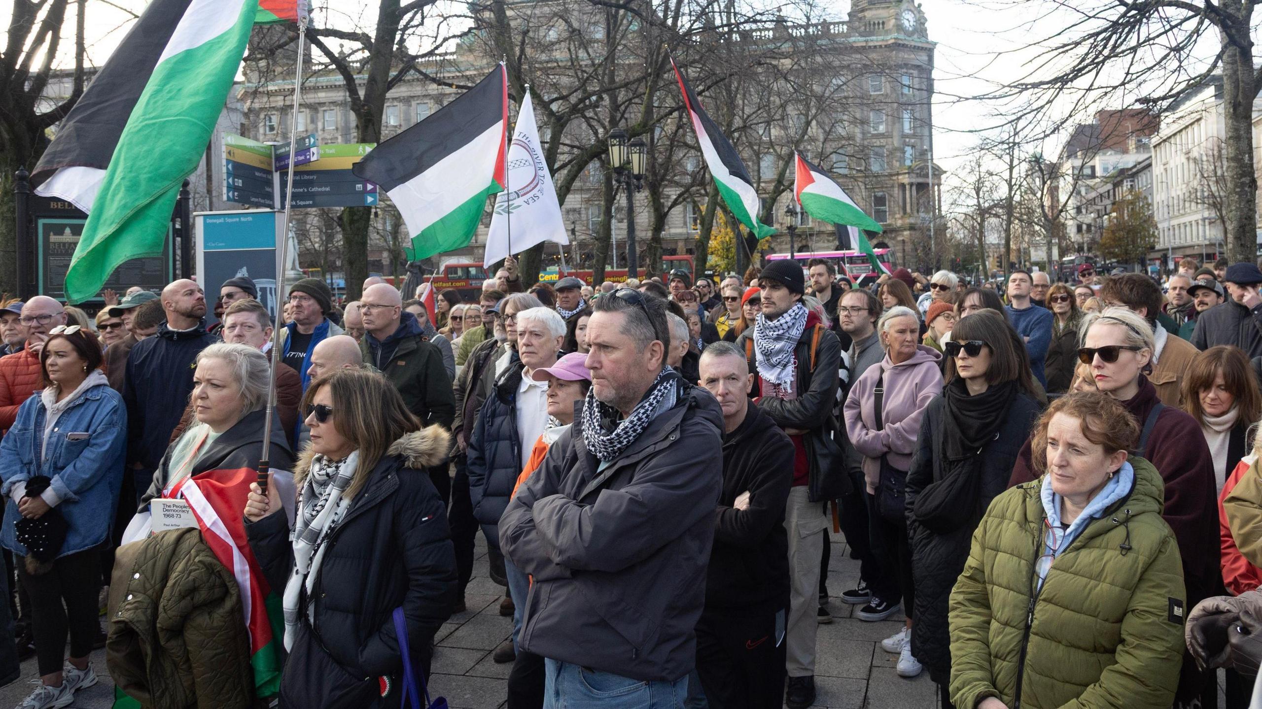 Protesters at the "Givan Must Go" rally.  A crowd of people stand listening to speakers in central Belfast.  They are wearing coats and scargs and some are holding Palestinian flags.  There are a number of tall buildings in the background.