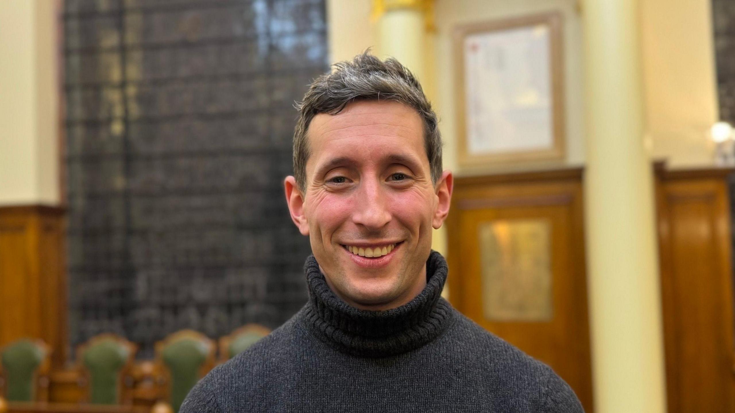 A man smiling straight at the camera. He is wearing a grey turtleneck jumper.