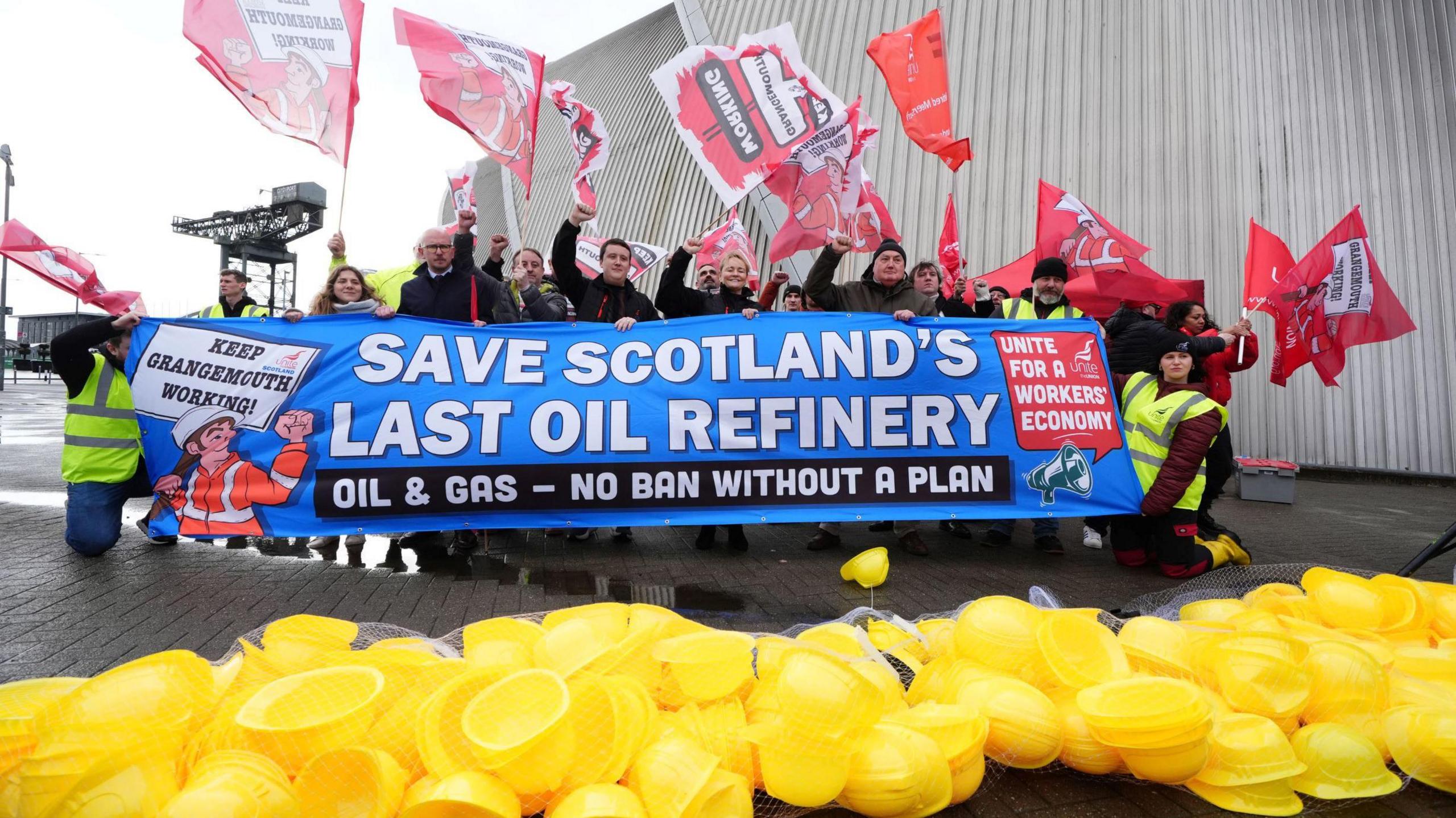 Unite union members protest - they are holding a banner saying SAVE SCOTLAND'S LAST OIL REFINERY up while waving red flags with slogans on them in the air. A large pile of yellow hard hats is in front of them.