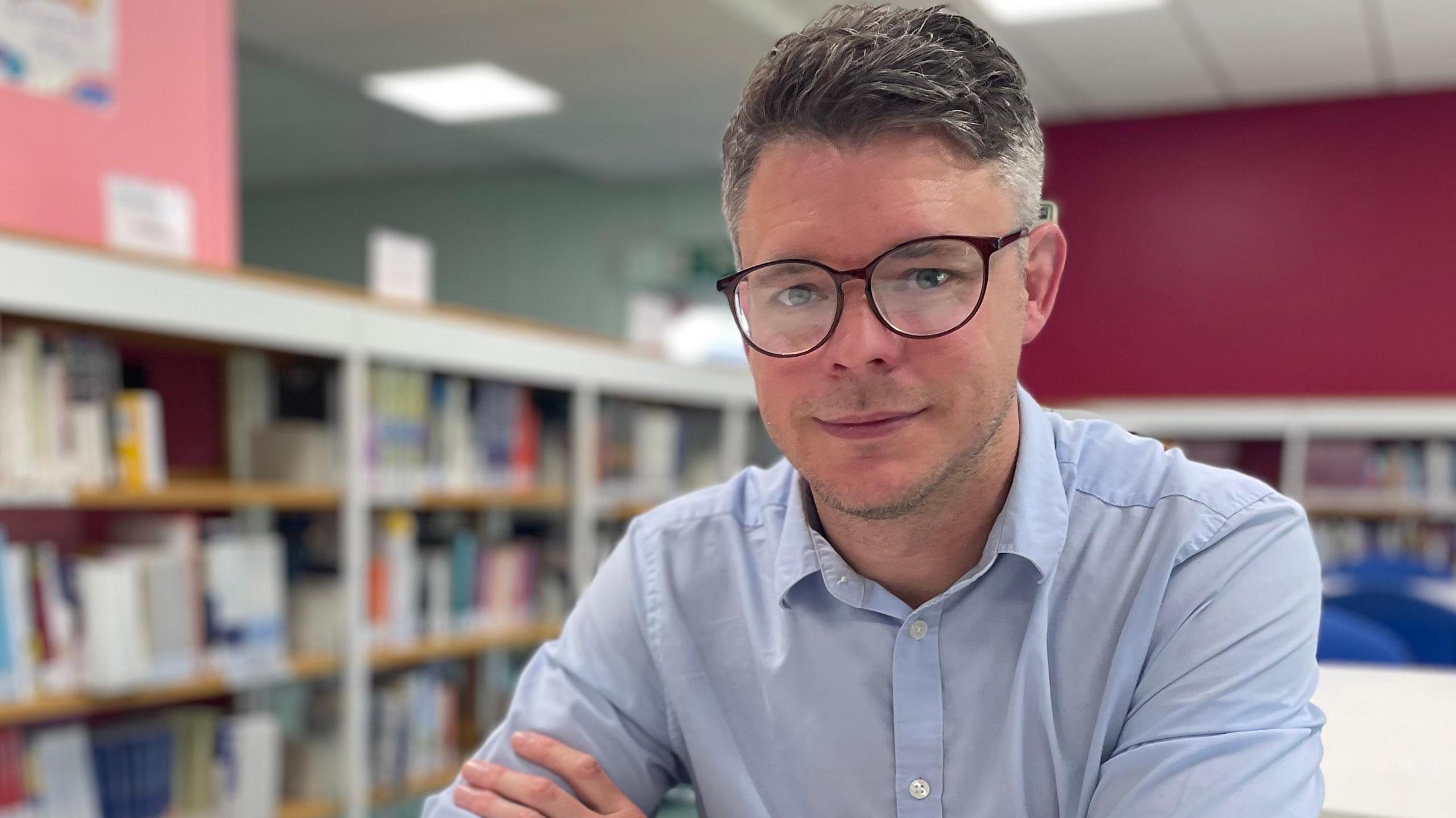 Teacher Rob Randel, wearing a blue shirt and brown rounded spectacles, looking at the camera in a library