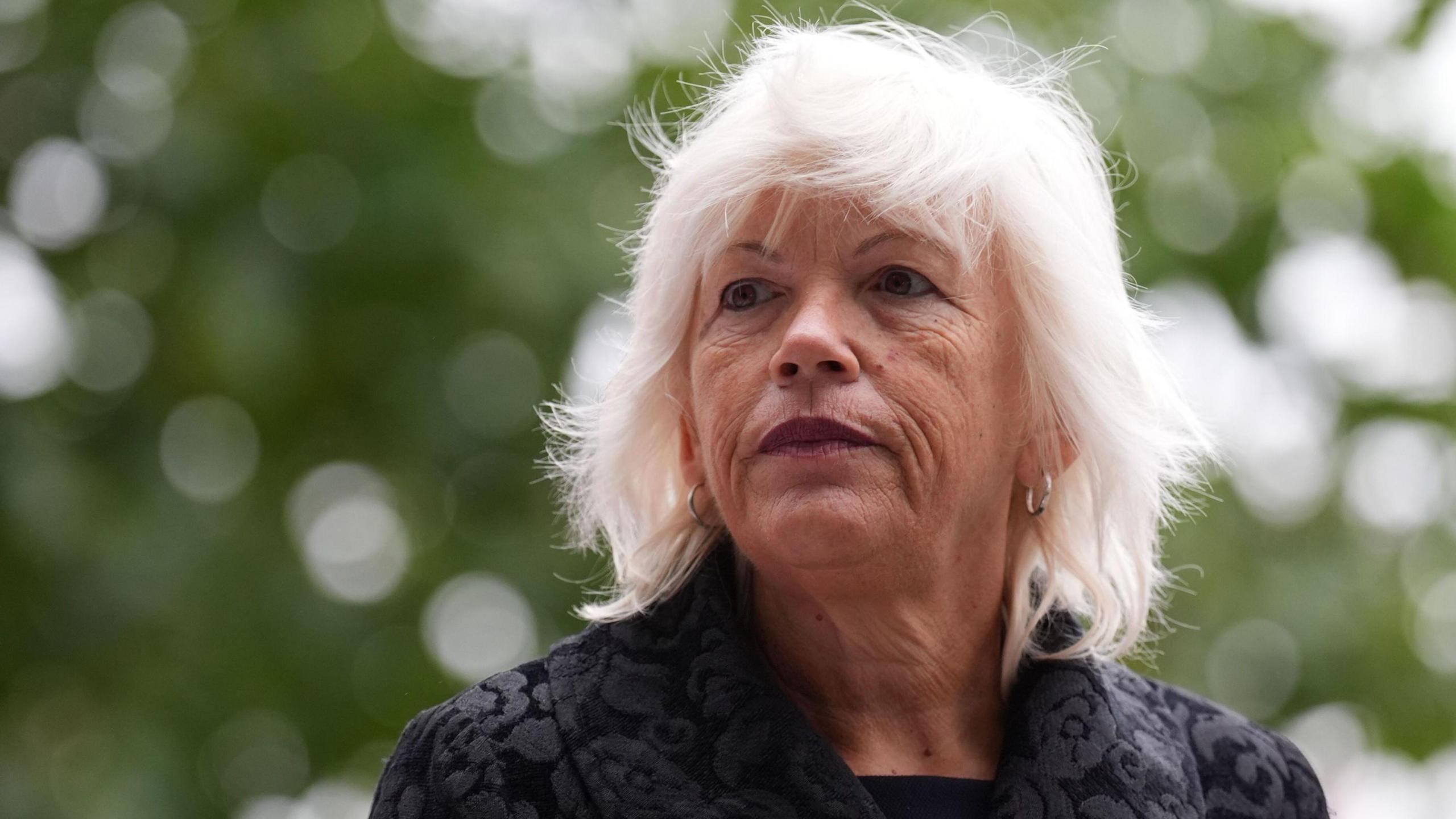 Melanie Leahy has white shoulder-length hair, and is wearing a dark jacket with floral pattern. She has small silver hoop earrings and wears dark red lipstick. She stands in front of a blurred background of trees