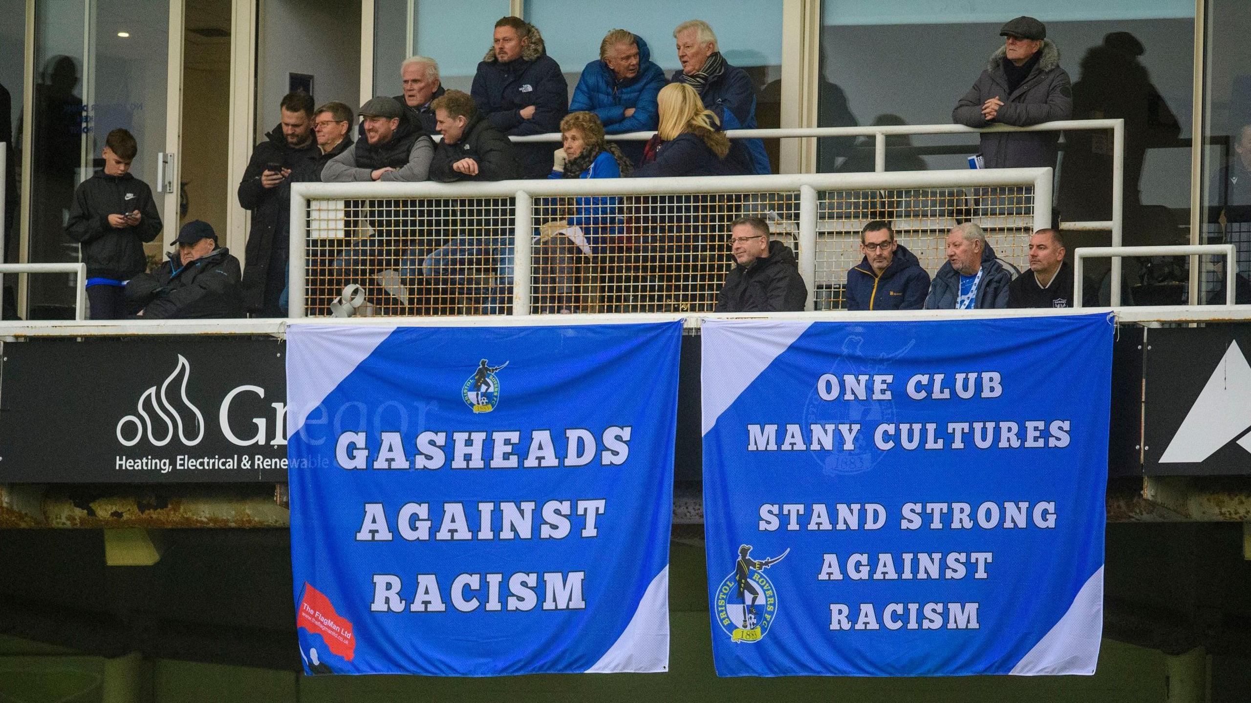 Two banners are seen hung over a barrier at the Memorial Stadium during Bristol Rovers' match with Lincoln City. One says "Gasheads against racism" and another says "one club, many cultures, stand strong against racism". Both banners are blue with the text in white.