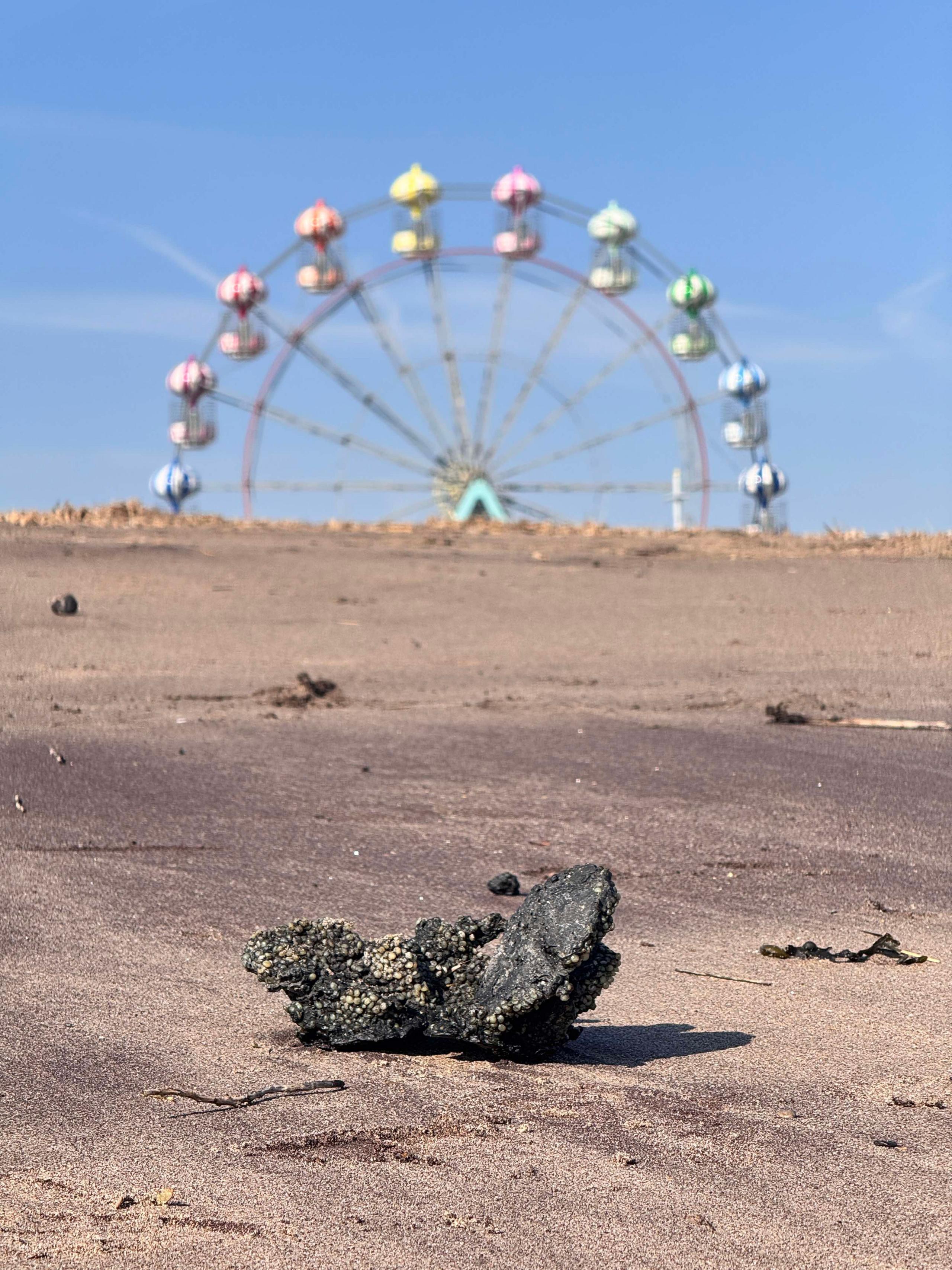 A burnt clump of what appears to be plastic pellets on the shoreline at Skegness with a big wheel in the background.