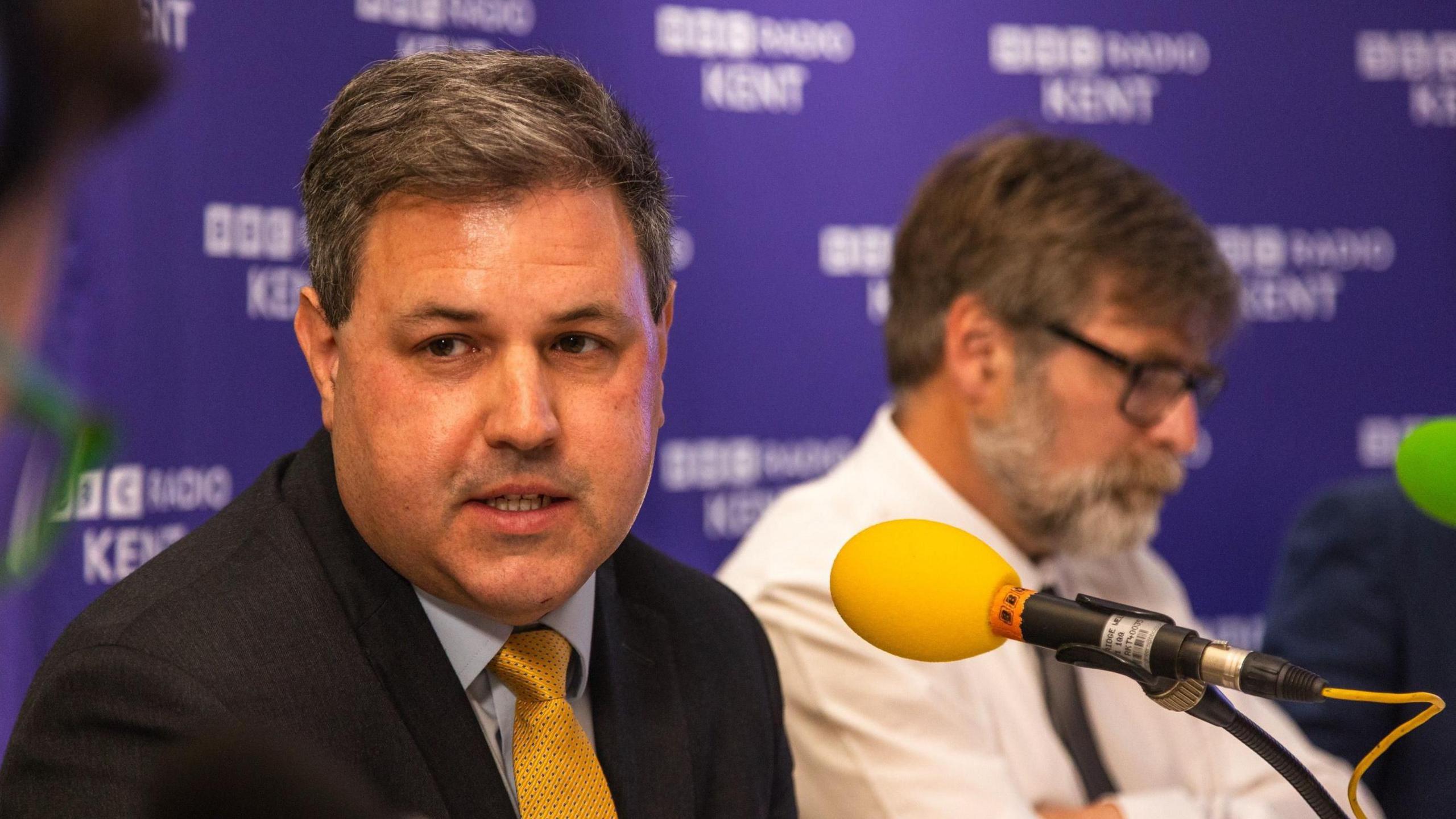 Antony Hook has dark hair, wears a dark suit, blue shirt and gold tie, as he speaks into a microphone during a BBC Radio Kent debate.
