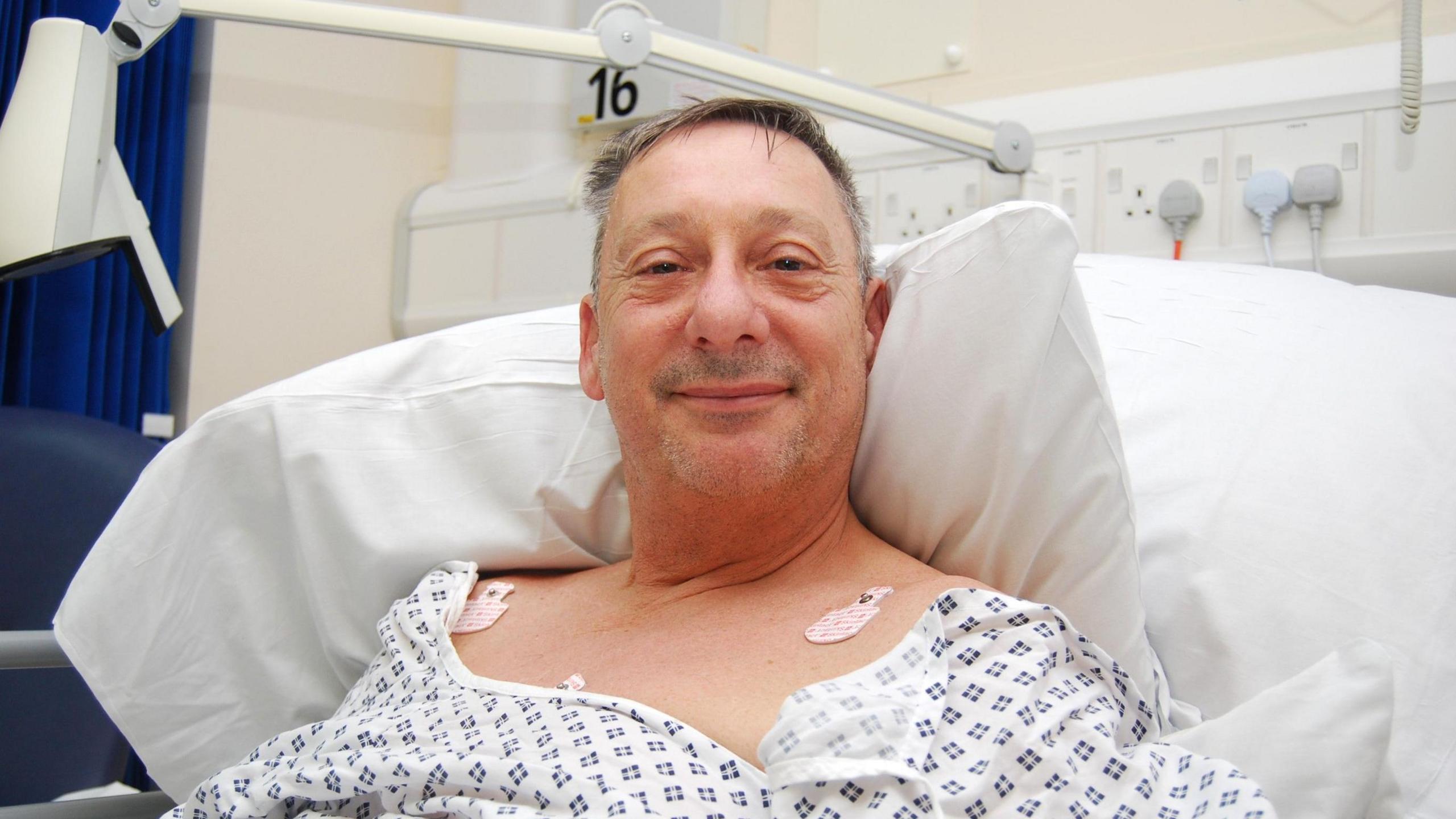 David McLoughlin laying on a hospital bed in a hospital gown. He is looking directly at the camera and smiling.