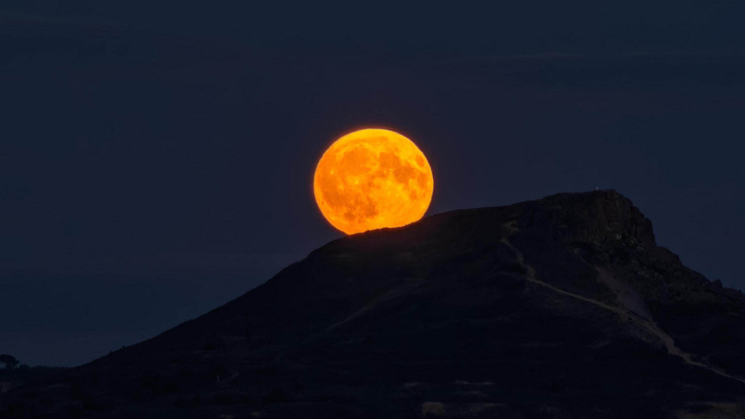 The circular orange moon is peeking out from behind the silhouette of the mountain. The sky is very dark blue.