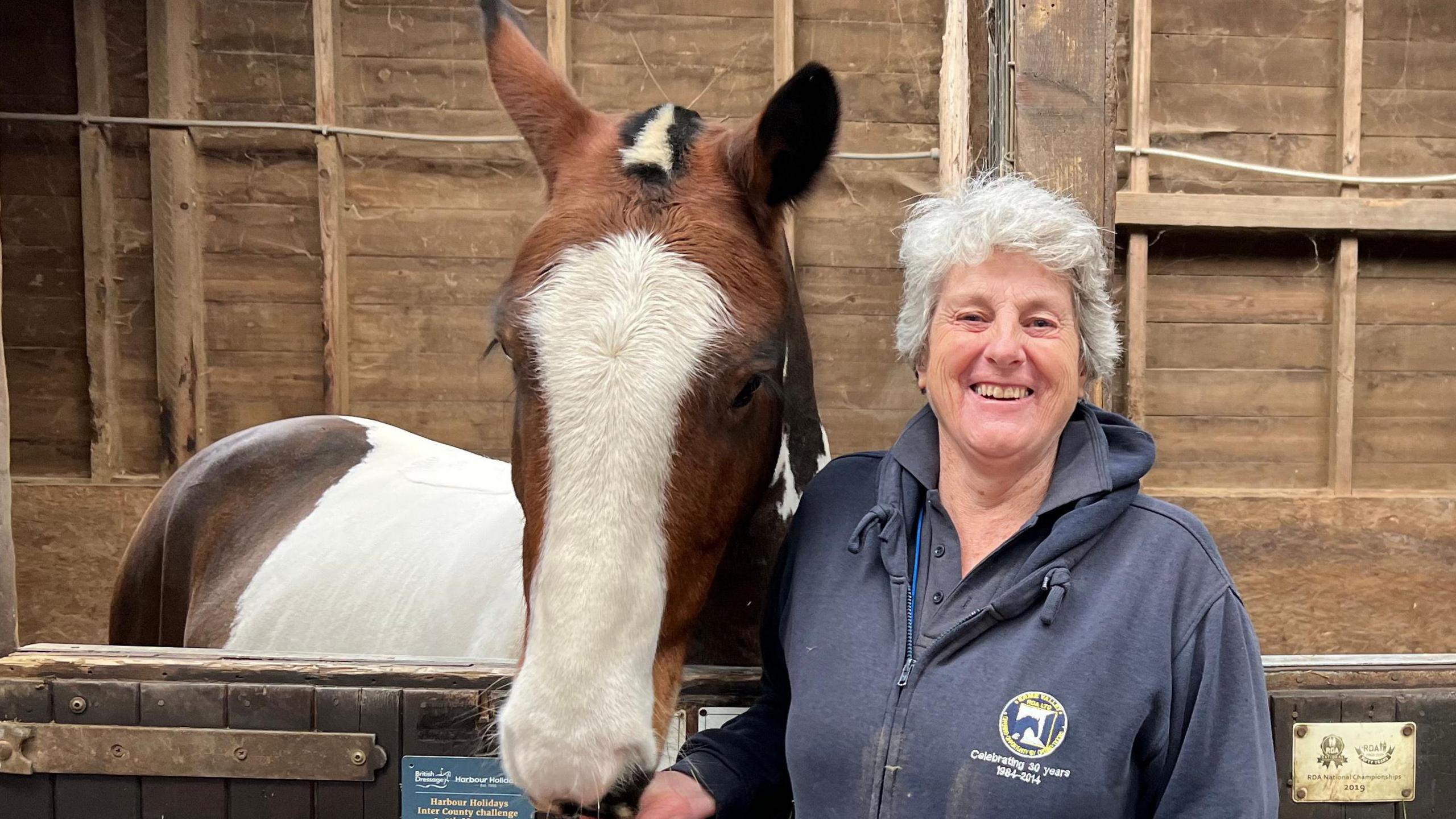 Peggy Douglas stood smiling at the camera and stood in a stable. She has her hand below a brown and white horse, which is taller than her. The barn is wooden and the horse is behind a gate with its head over the top.