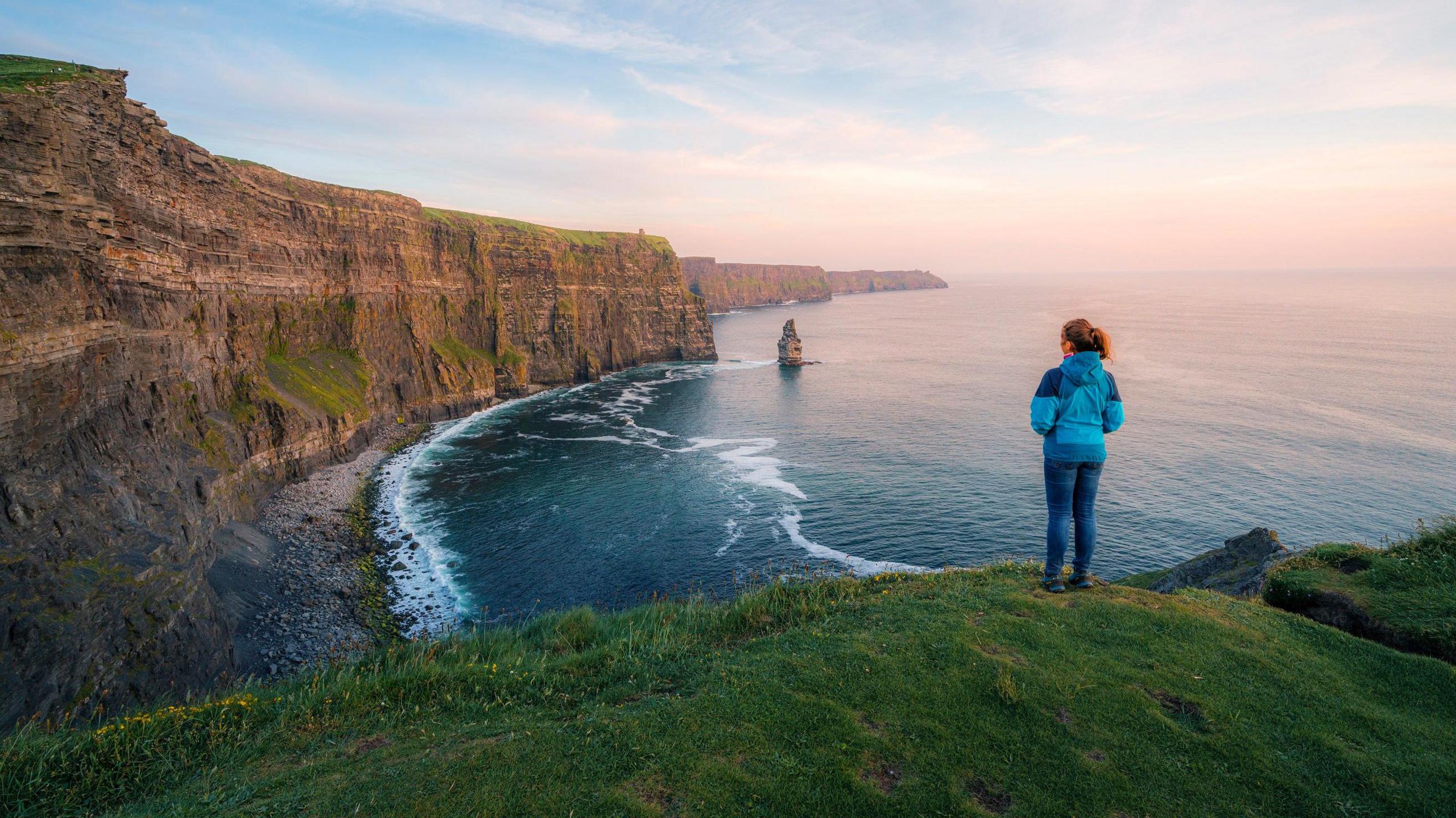 A woman stands on top of a cliff gazing at the Cliffs of Moher at sunset in late spring. She is wearing a blue coat and jeans, the sea is below her, the cliffs in front.