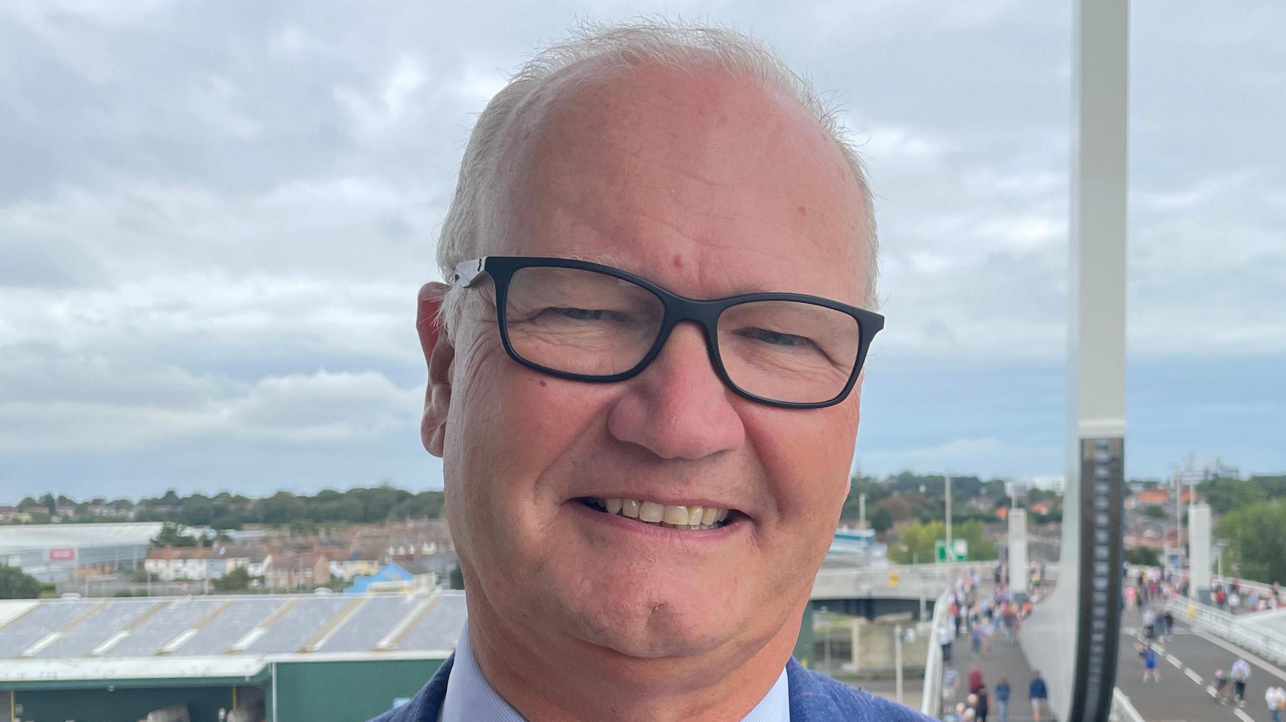 A man with thin grey hair smiles at the camera. He is wearing black glasses with a blue suit and blue shirt. Behind him a bridge is pictured with people walking across it.