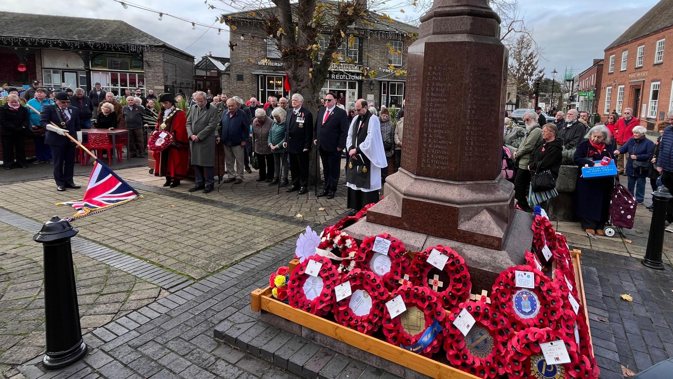 A large crowd of people surrounding a war memorial, which has poppy wreaths at its base. A man is holding a Union Jack flag which he has lowered to the floor.
