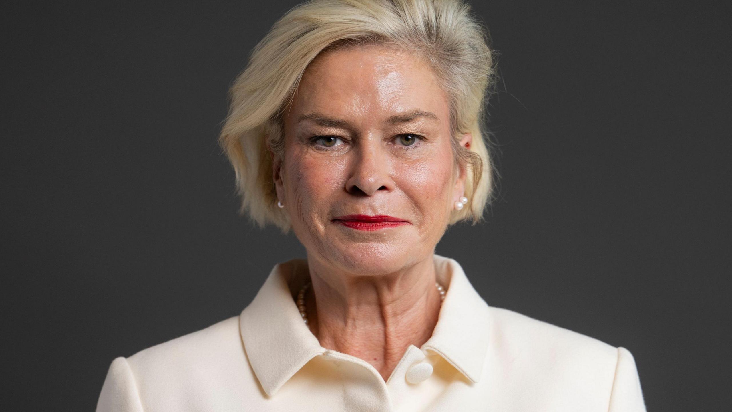 An official portrait photo of the MP for Taunton Rachel Gilmour. She has blonde hair and is wearing a cream jacket. The picture is taken against a grey background