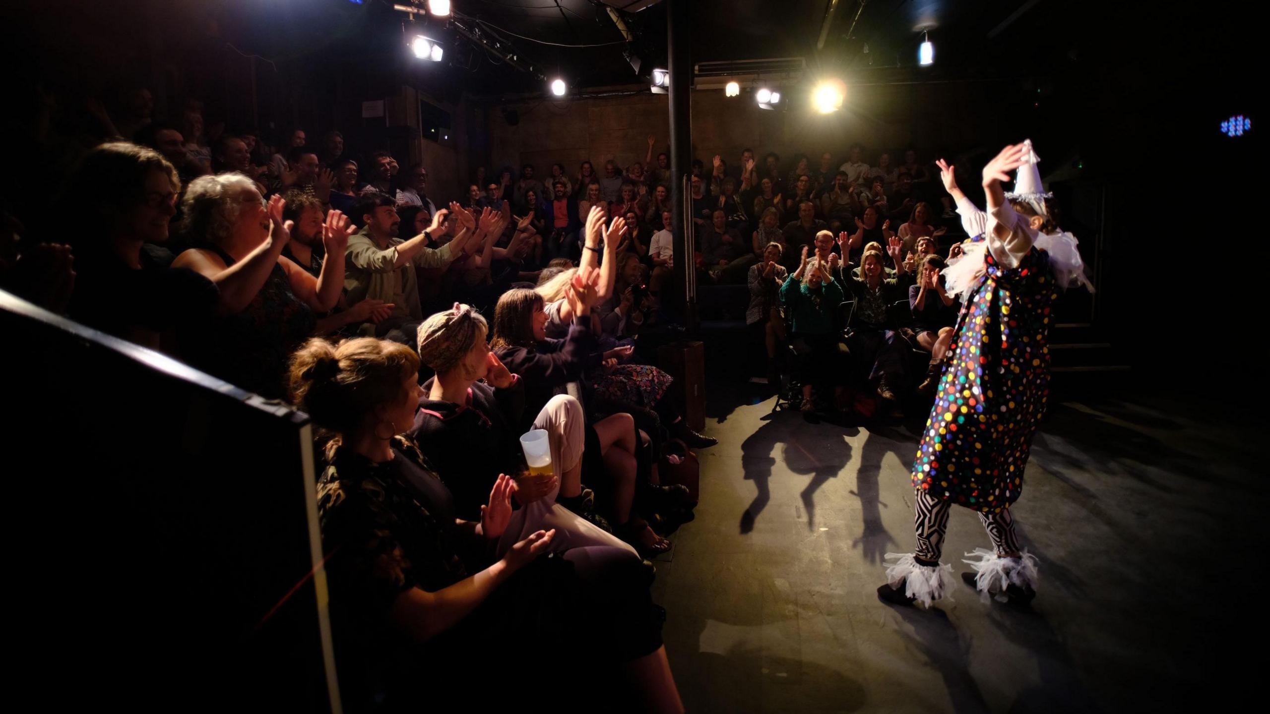 A performer in a polka dot dress and striped trousers takes the applause of the audience at the Wardrobe Theatre in Bristol. The venue is dark but there are spotlights shining on the stage