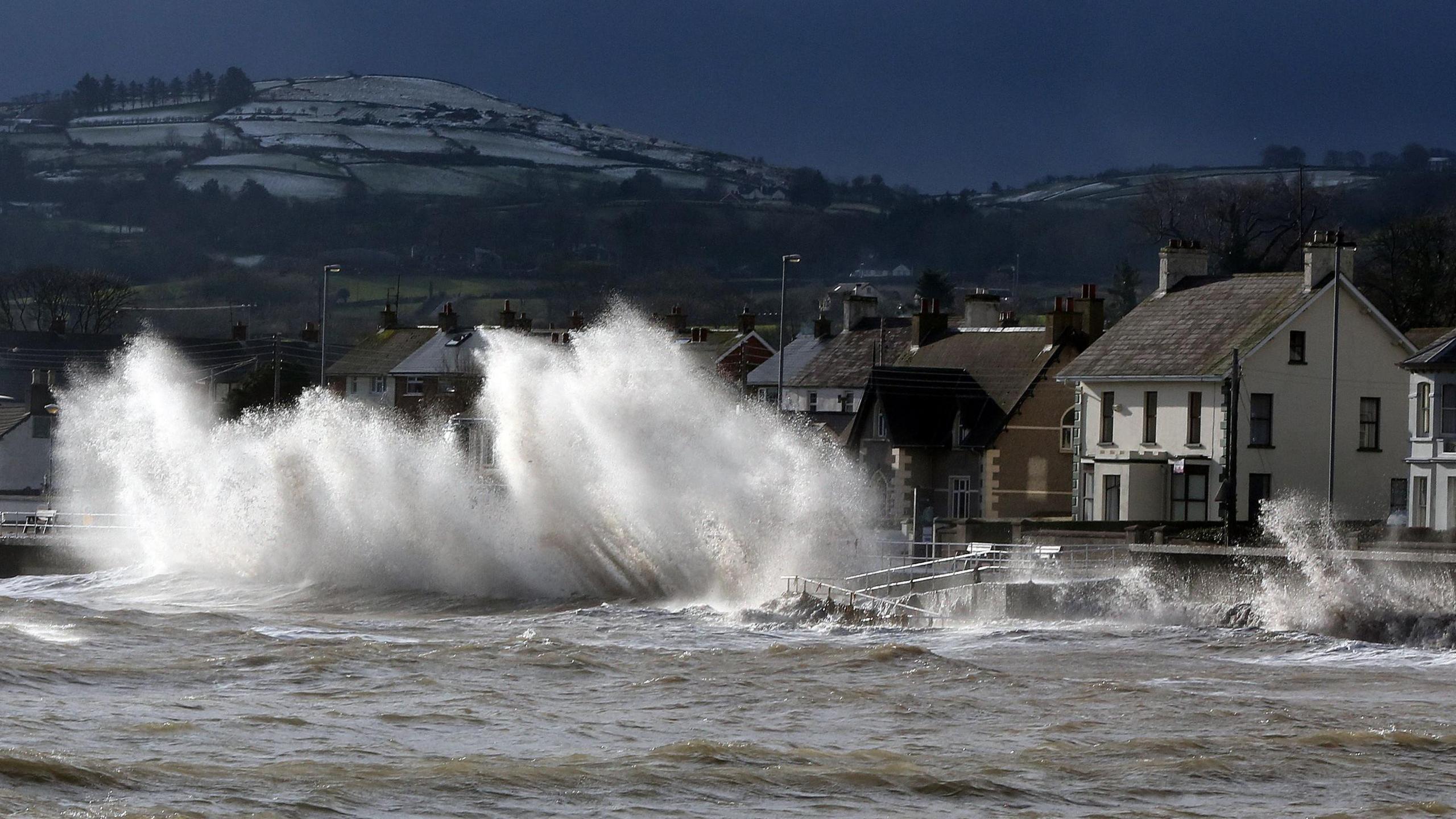 Large white waves crash against the road during a storm. There are a number of visible houses. In the background there are hills covered in snow.