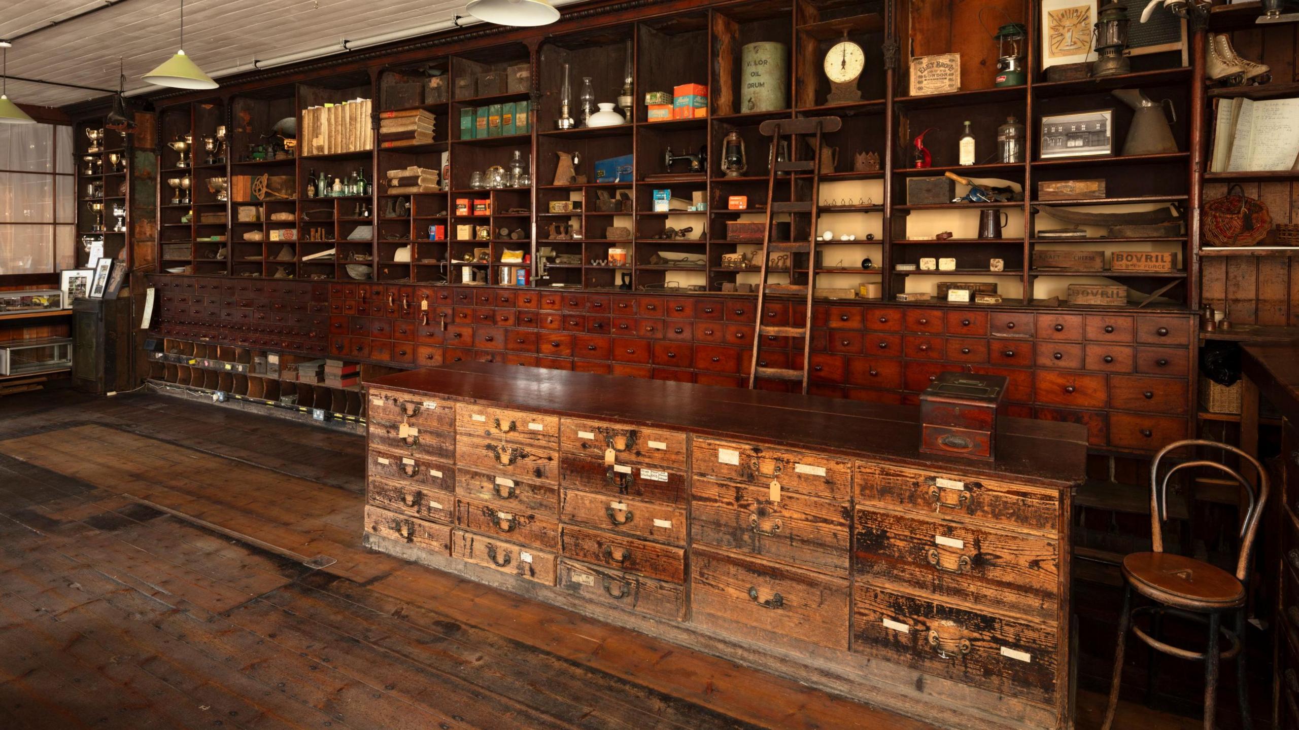 The large shop interior has wooden shelving and drawers across its back wall, a wooden counter with large drawers and a wooden chair to the side.