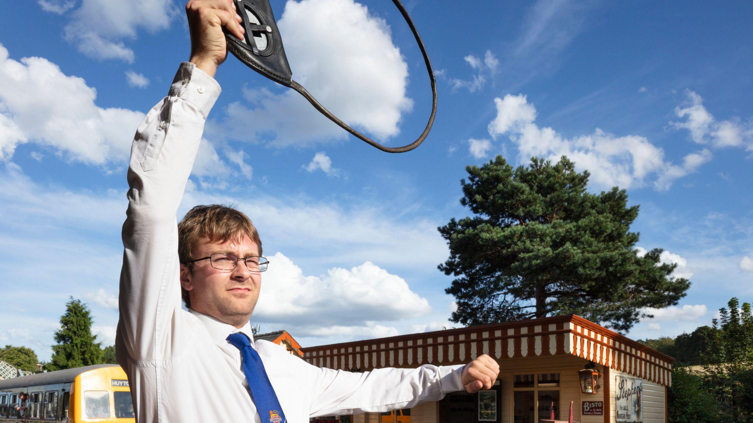 A man holds a circular rail signal above his head with his right hand as he stands next to a rail line in a white shirt and blue tie. A train carriage has stopped at the platform behind him.