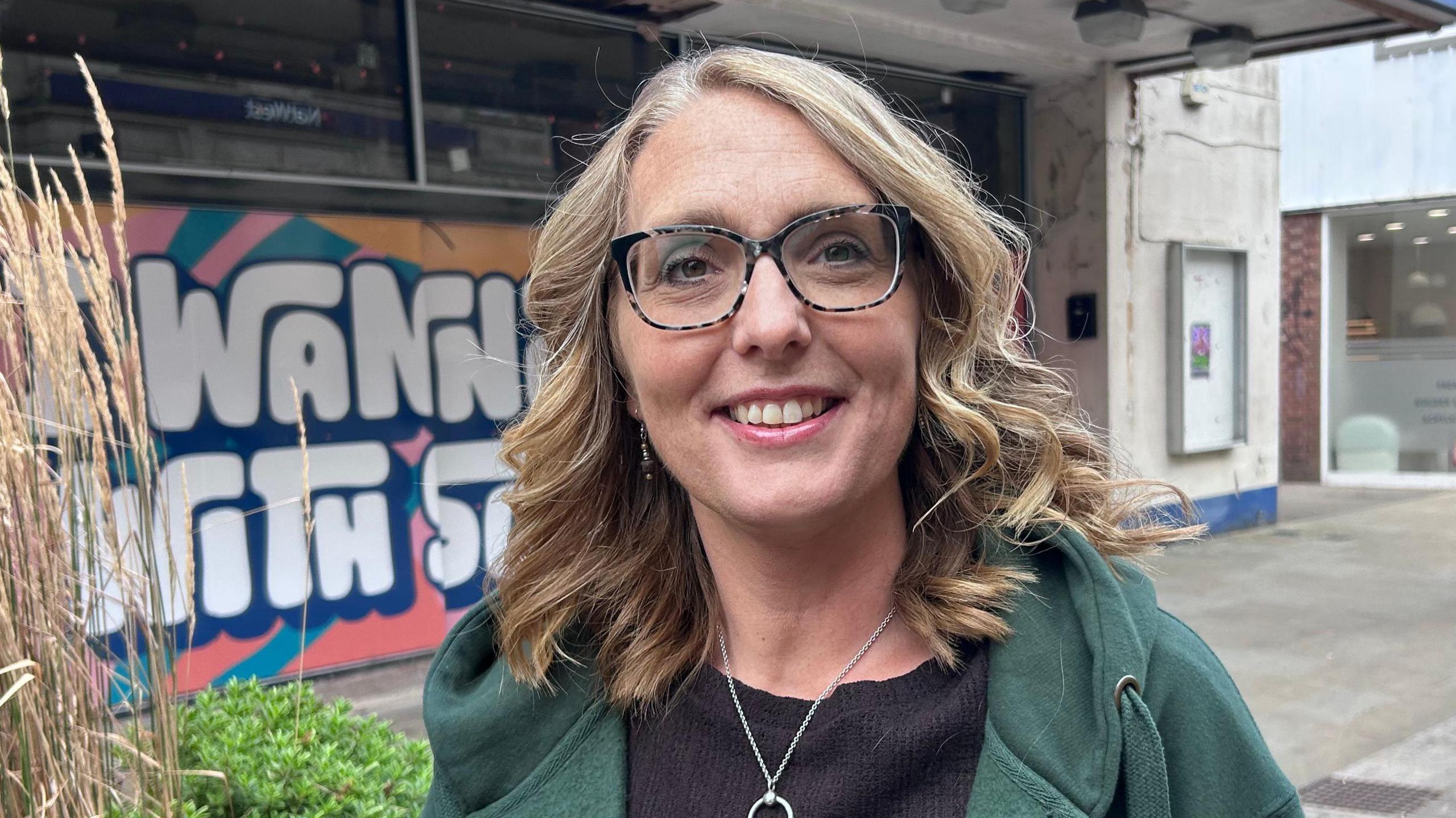Lindsey Atkins standing in Kettering high street. She is in a green hooded jumper and wears eye glasses with large frames. Her hair is shoulder length and blonde and she is smiling at the camera.
