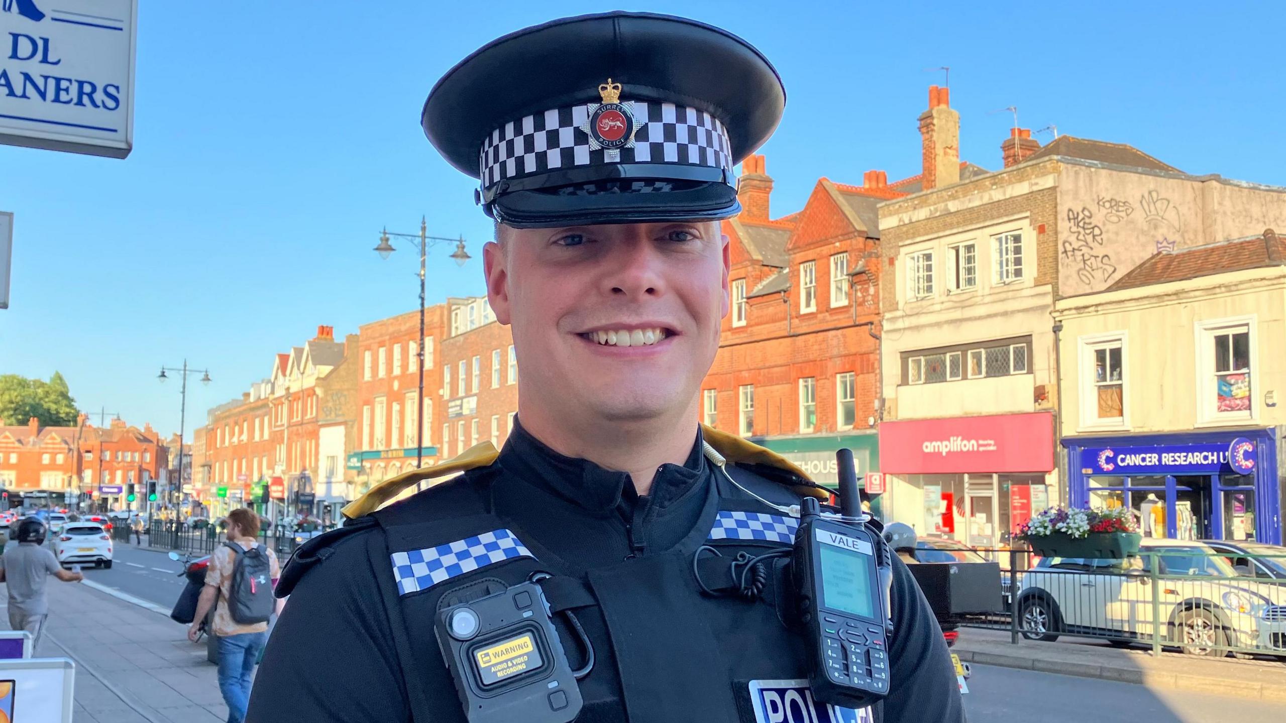 Surrey Police Inspector Jon Vale looks at the camera and smiles. He is wearing a police uniform and hat and in the background is Epsom High Street in the evening sunshine.
