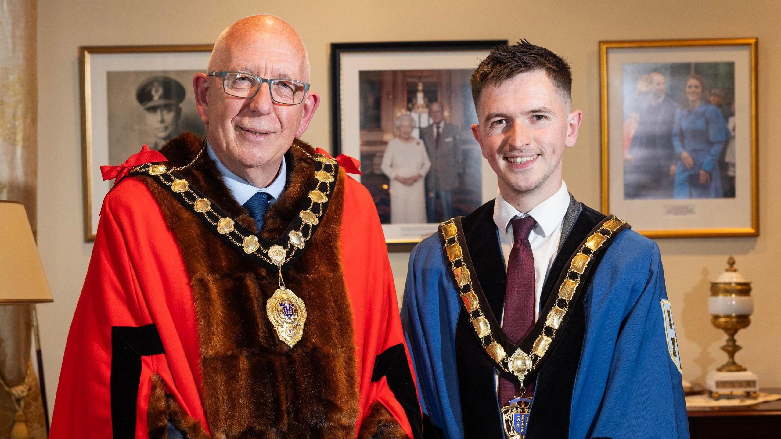 Mayor Jackson Minford and Deputy Mayor Tyler Hoey pictured smiling at the camera in an office.  Minford is wearing a red, fur-trimmed ceremonial robe over a grey suit, with the gold chain of office around his neck.  He has grey-rimmed glasses and no hair.  Hoey has short, dark hair and is wearing a blue ceremonial robe and chain over a grey suit with a white shirt and burgundy tie. 