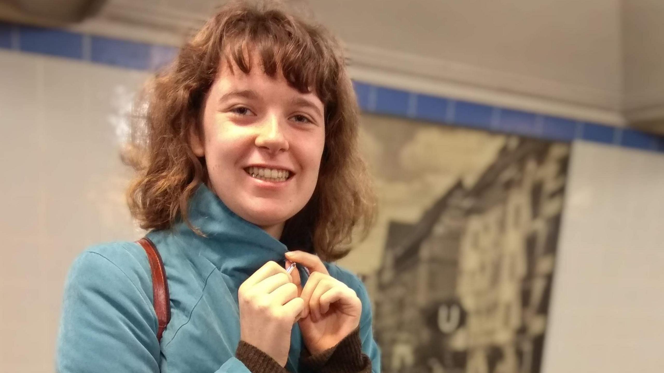 A young woman with shoulder length brown hair smiled for the camera as she holds the zip on the collar of her green jacket. She appears to be standing in a brightly lit tunnel with a black and white mural partially hidden in the background. 