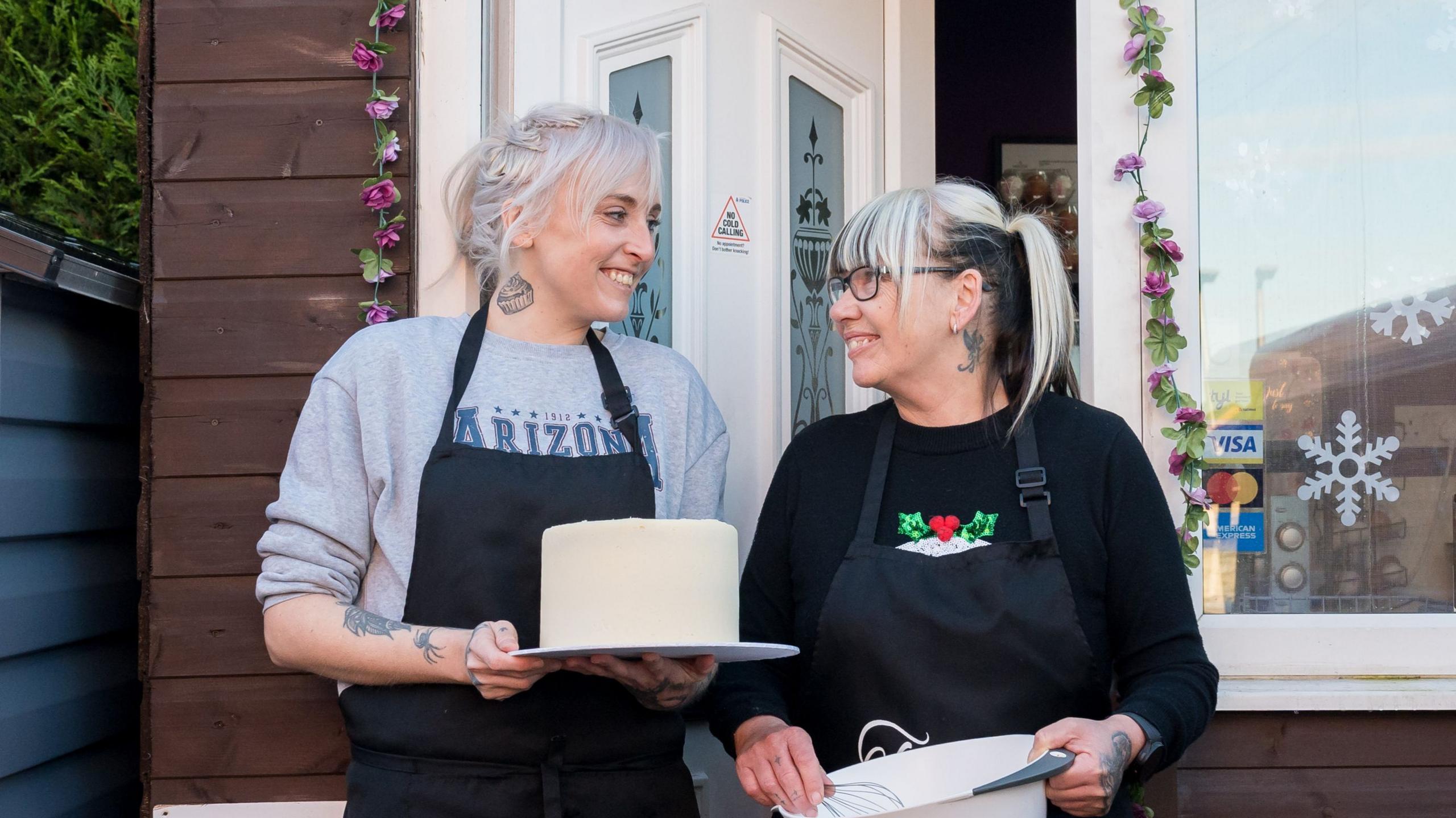 Two blonde women look at each other, smiling. They are standing outside what looks like a hut or wooden building, but what is actually a converted shipping container. They have aprons on, and are holding a cake and baking utensils.