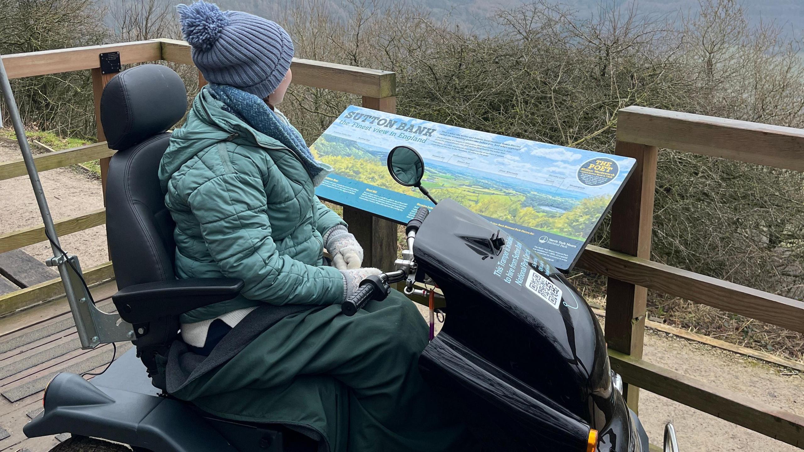 A young woman with a woolly hat sits on a large black off-road mobility scooter. She wears a green puffed jacket and a blue and white striped jumper. She is parked on a raised wooden viewing platform overlooking a countryside scene.