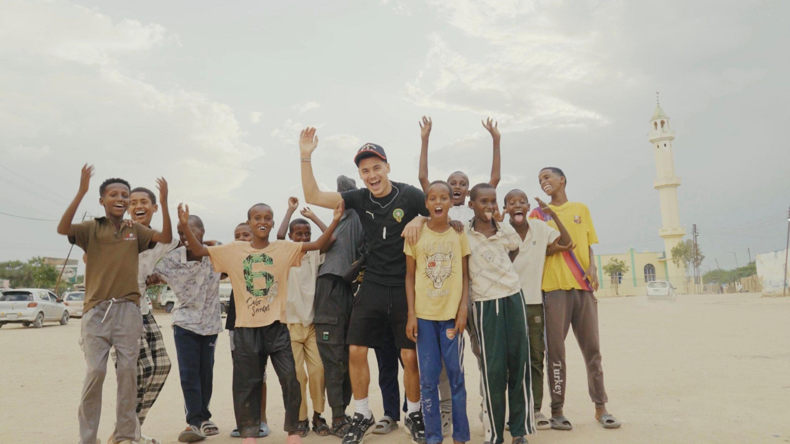 Zak Hajjaj in Somaliland standing in the middle of a bunch of local Somali kids outside, all smiling waving their hands