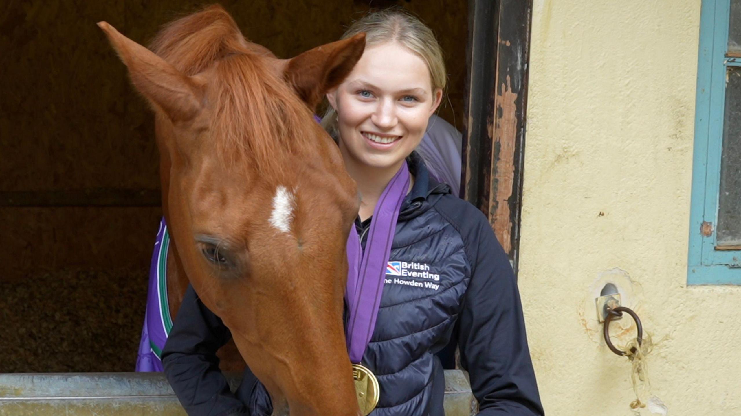 Ella smile at the camera in front of a stable door. She has blonde hair and wears a gold medal around her neck. She is also wearing a navy puffer coat. Spice the horse rests his head over the stable door and nuzzles her hand. He is a chestnut coloured horse with a speck of white at the top of his head.