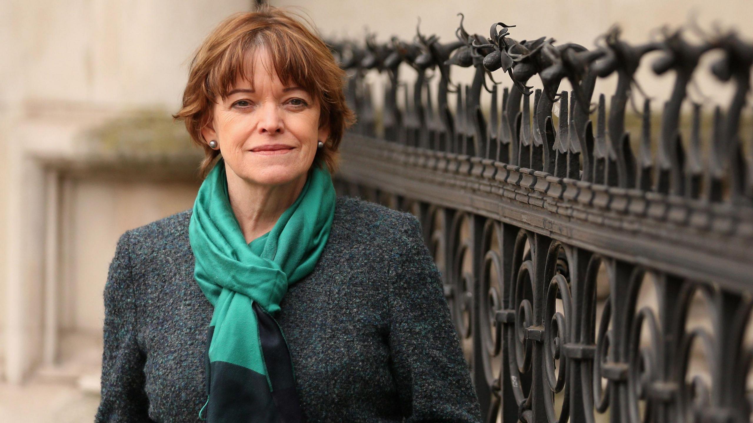 Dame Glenys is standing outside a court beside black fencing in the shape of flowers. She's wearing a grey coat and a green scarf.