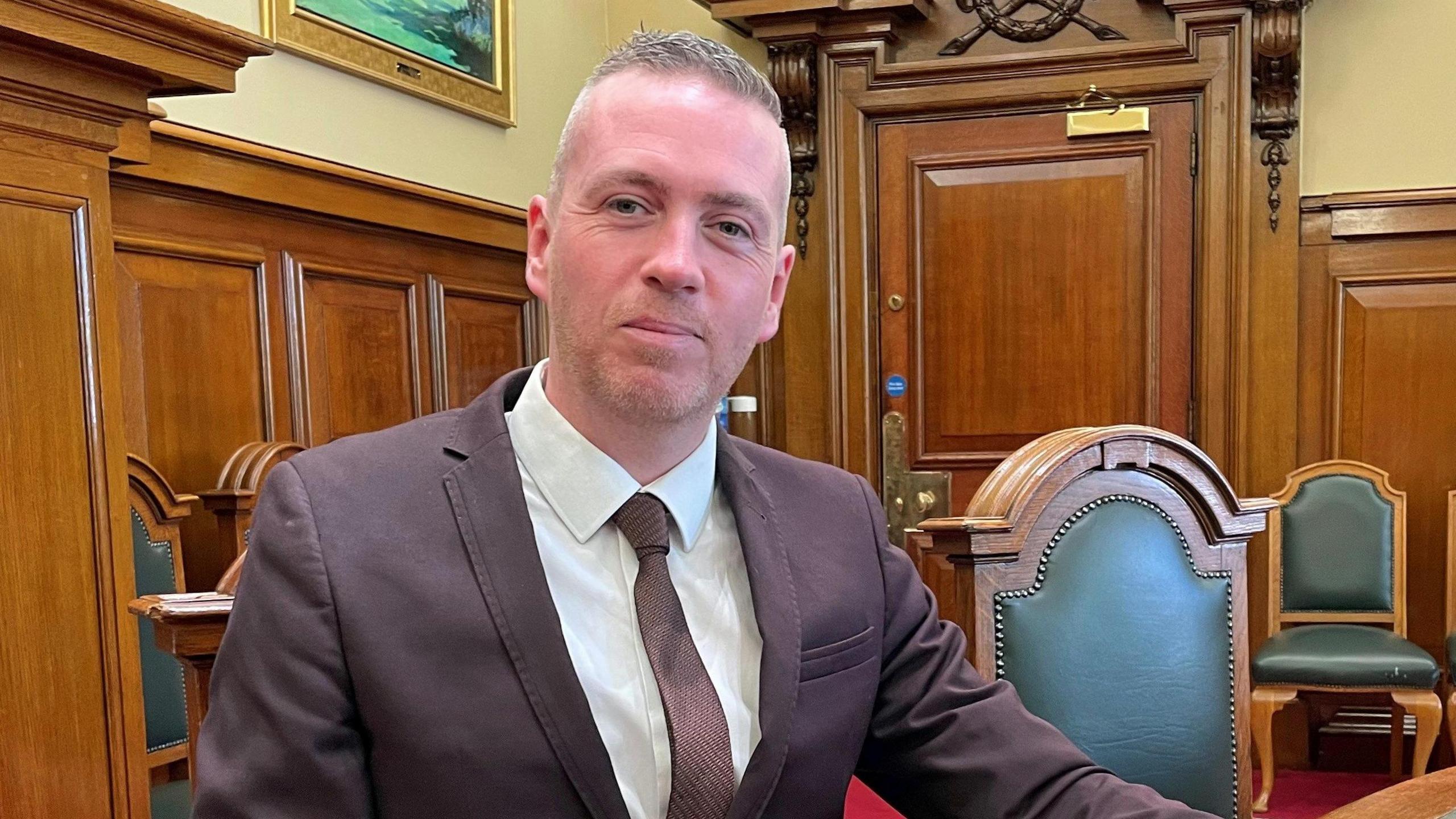 O'Hara is wearing a suit and a tie. He's sitting in an official looking room, with chairs and wood panelling. 