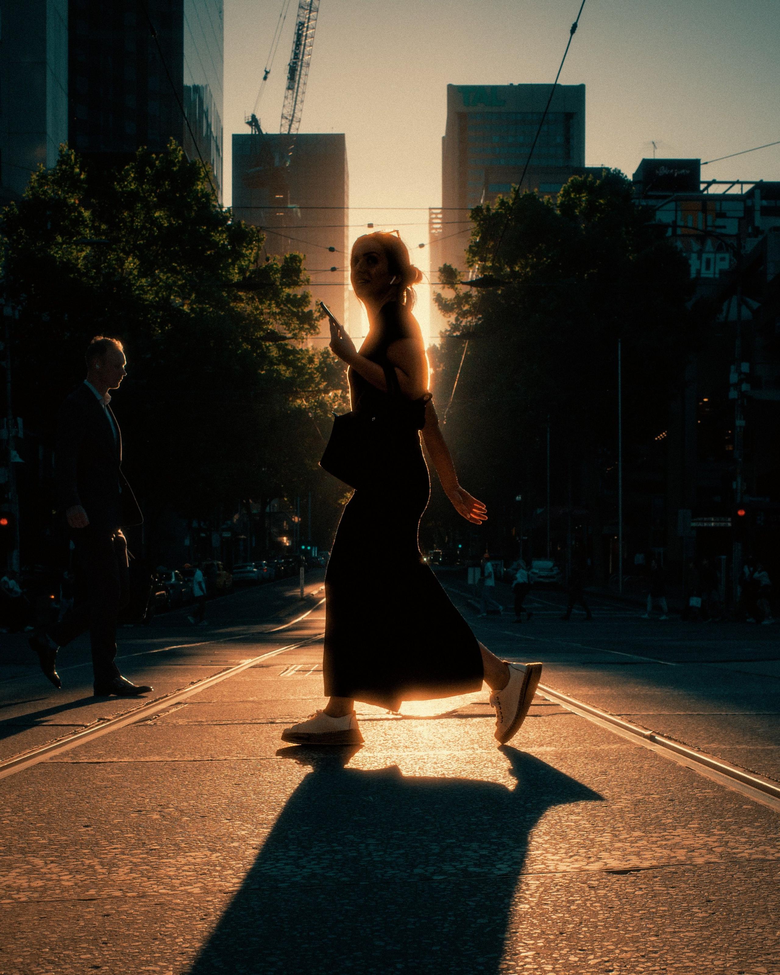 A woman silhouetted against the sun. She is striding across a road. There are skyscrapers in the background.