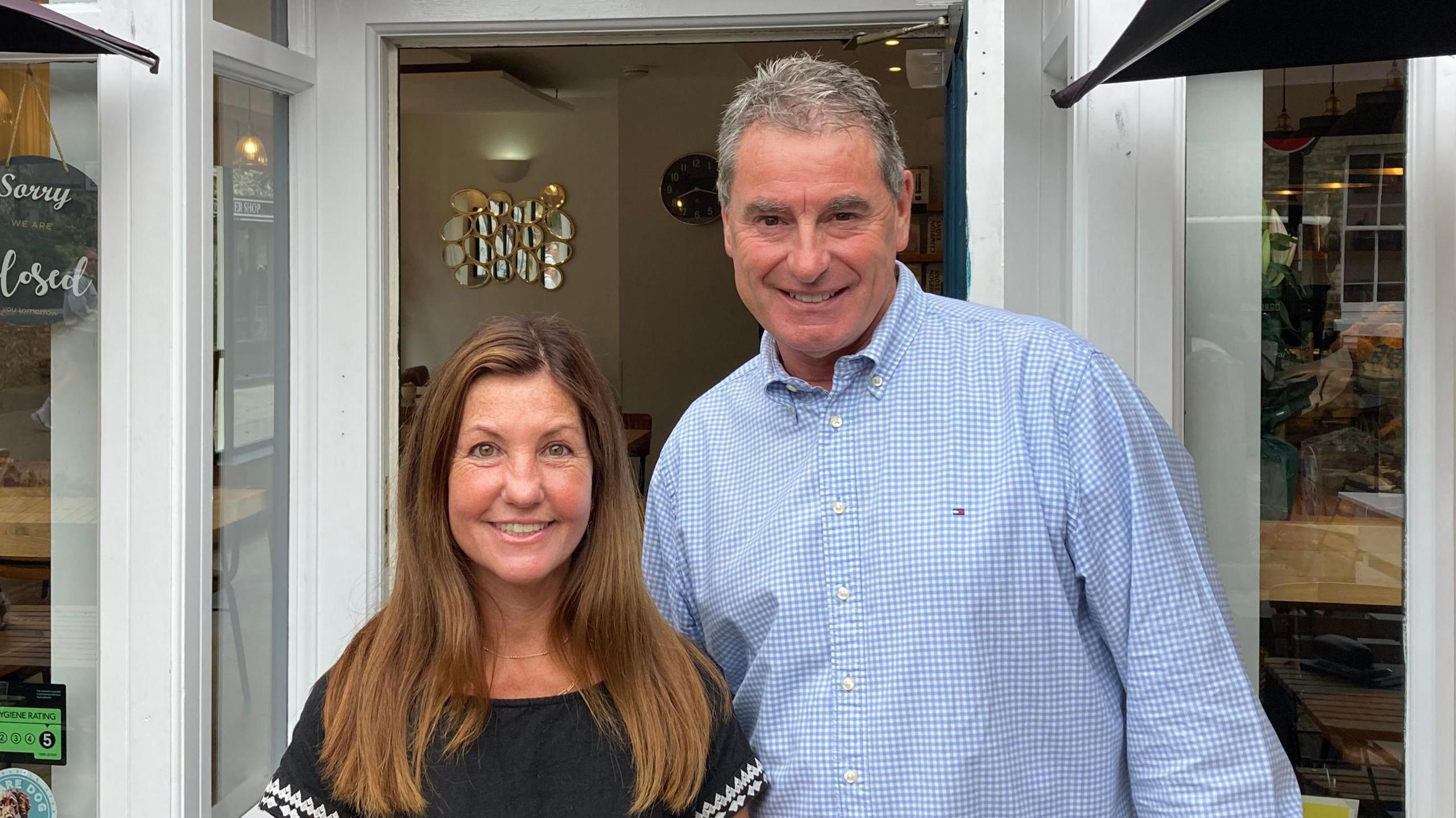 Steve Elsom and Vicky Snell stand outside a coffee shop and smile. Vicky has long brown hair and wears a navy top. Steve has short grey hair and wears a blue shirt.