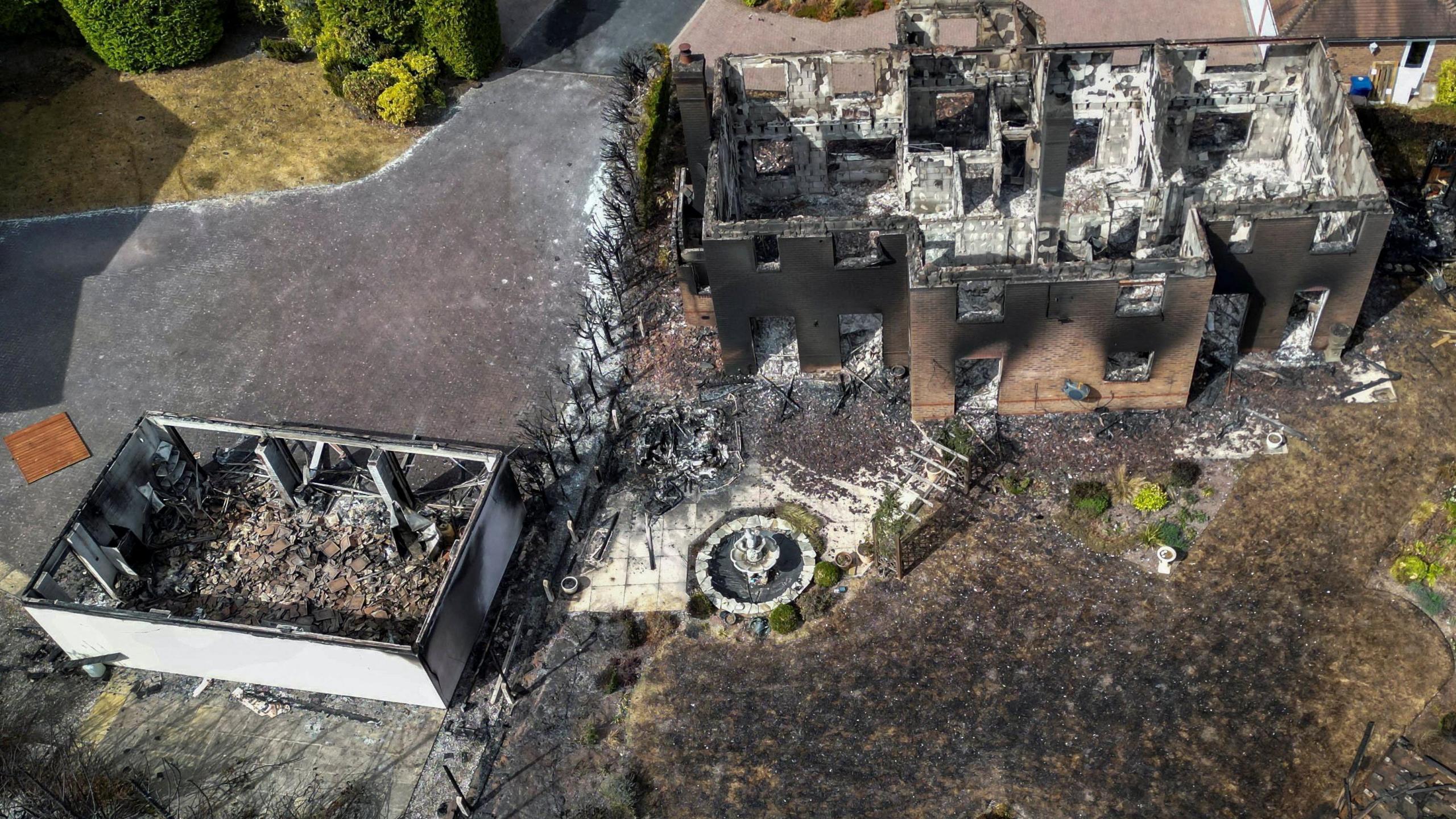 An aerial photo of a large house and garage next to it completely destroyed by fire. There are no roofs and doors on either building and they are black and charred from fire. The insides are rubble and white