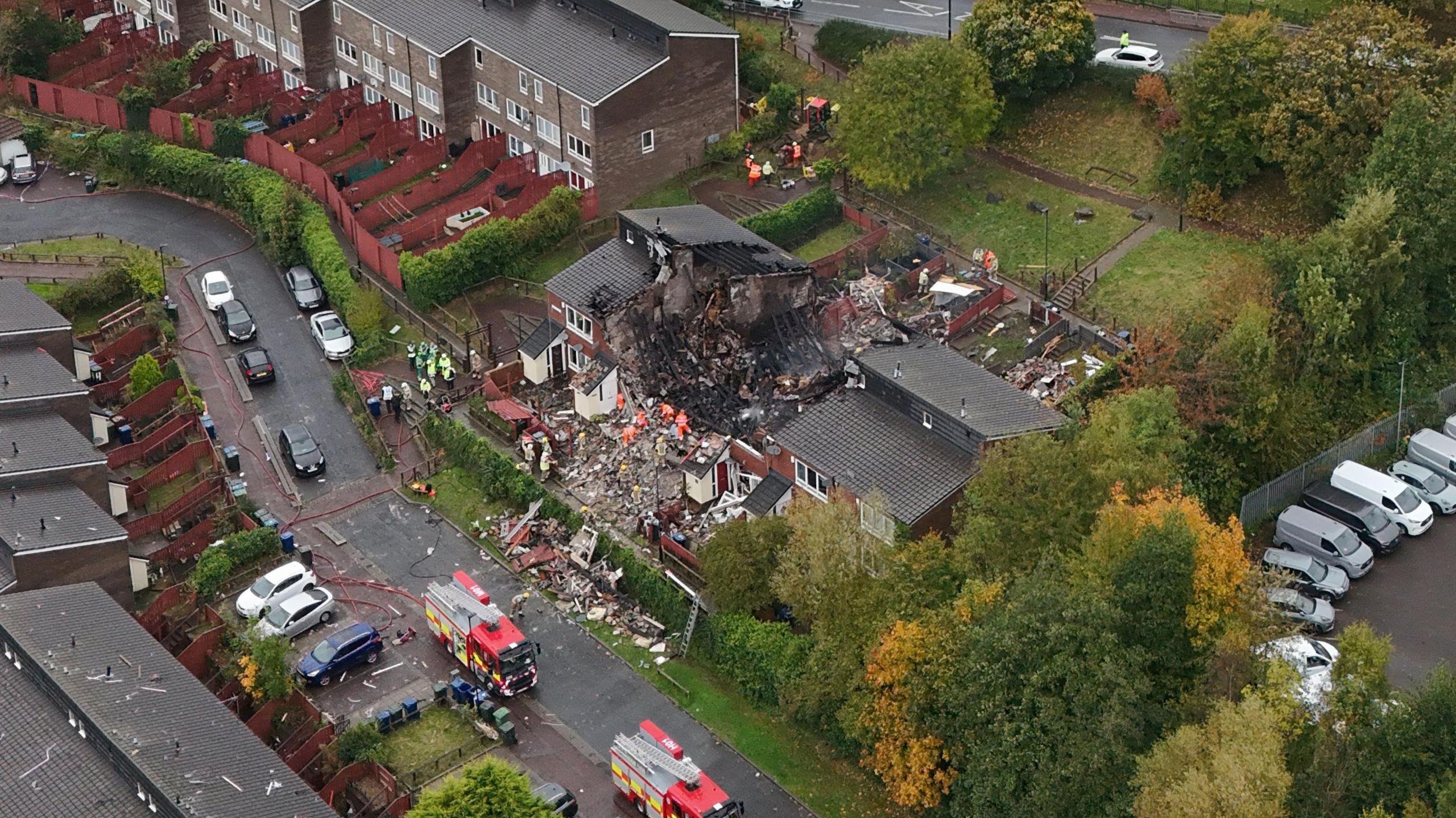 Aerial shot of damaged properties. One home's roof has been destroyed. Debris from the building is scattered on the nearby grass and street. Two fire engines are parked on the road and firefighters are clearing rubble from the building.