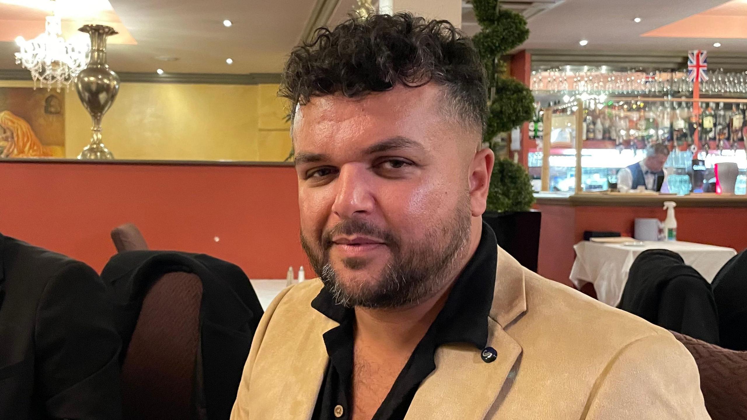 Alex Rafiq, looking direct to camera, seated in a restaurant. He has curly, black hair, cut shorter at the sides and longer on top, and dark eyes. He is wearing a camel coloured jacket and black shirt, open at the collar. Over his shoulder on the right of the image is a bar area where a member of staff can be seen working. 