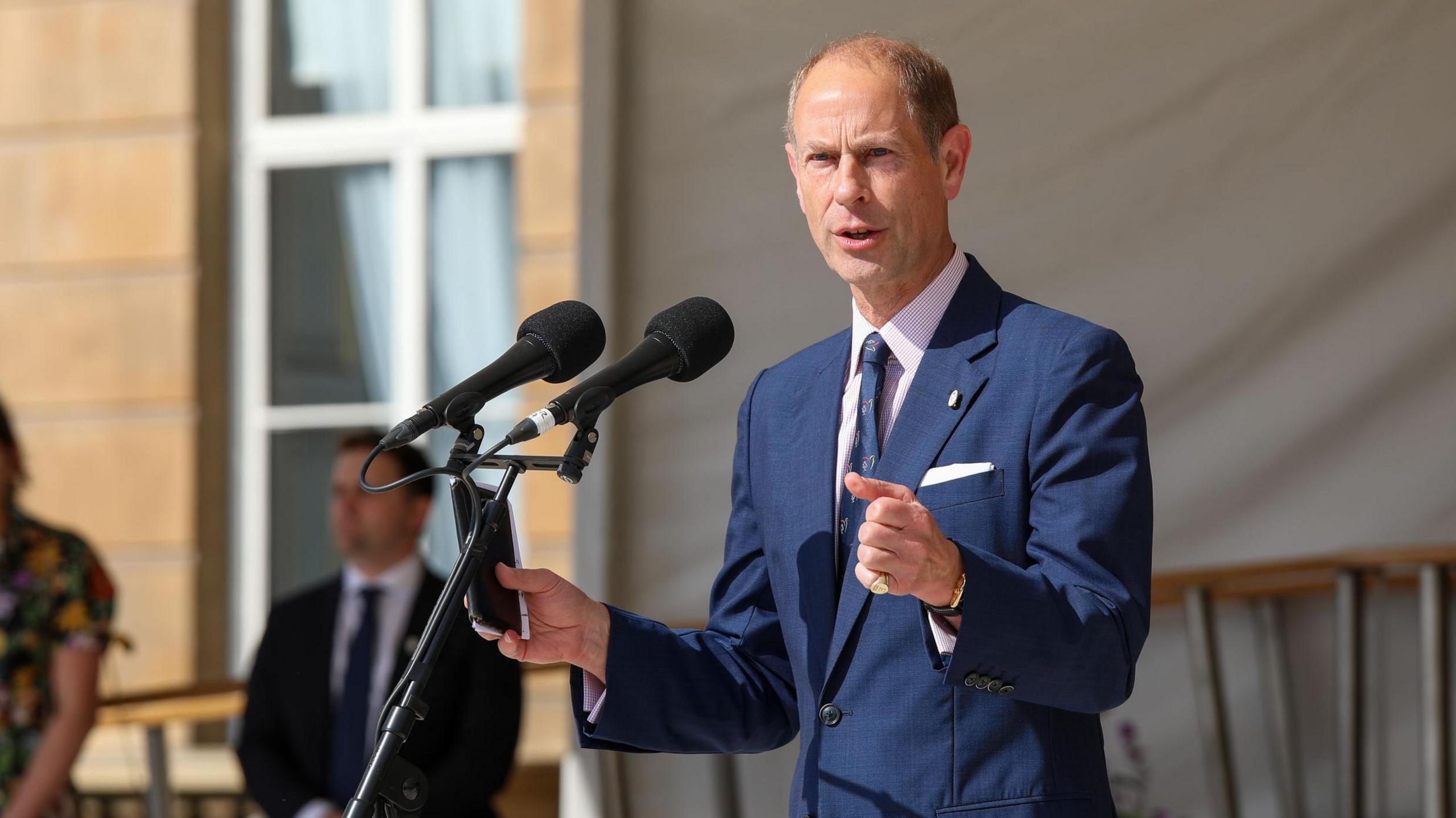 Prince Edward with short grey hair, wearing a blue jacket and tie, holding his hands forward and speaking into two microphones on a stand. There is a man in a suit standing in the background and the light stone of Buckingham Palace is visible behind.