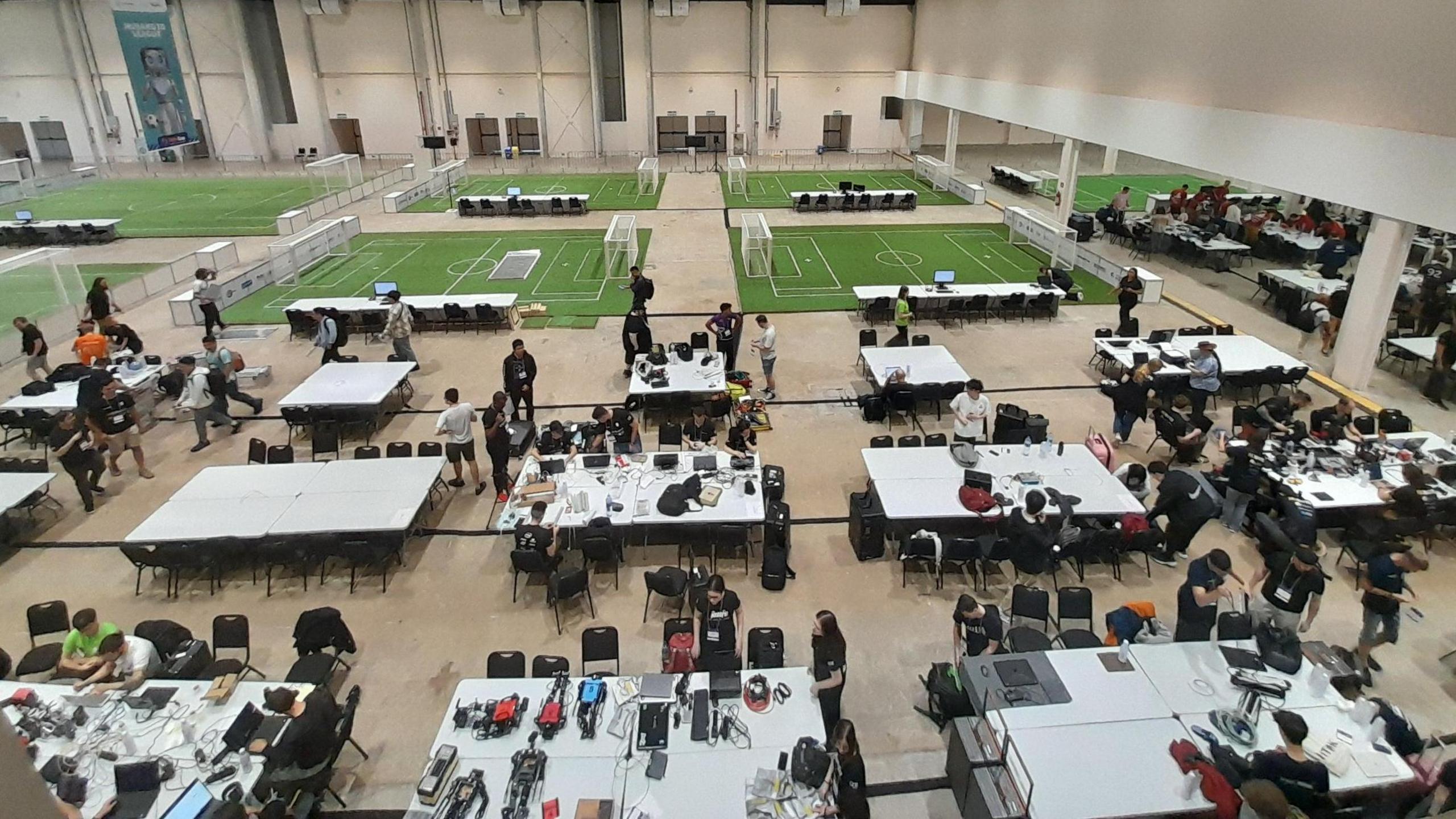 An aerial view of a large arena which has several mini football pitches in it. There are also large tables with teams of people around them.
