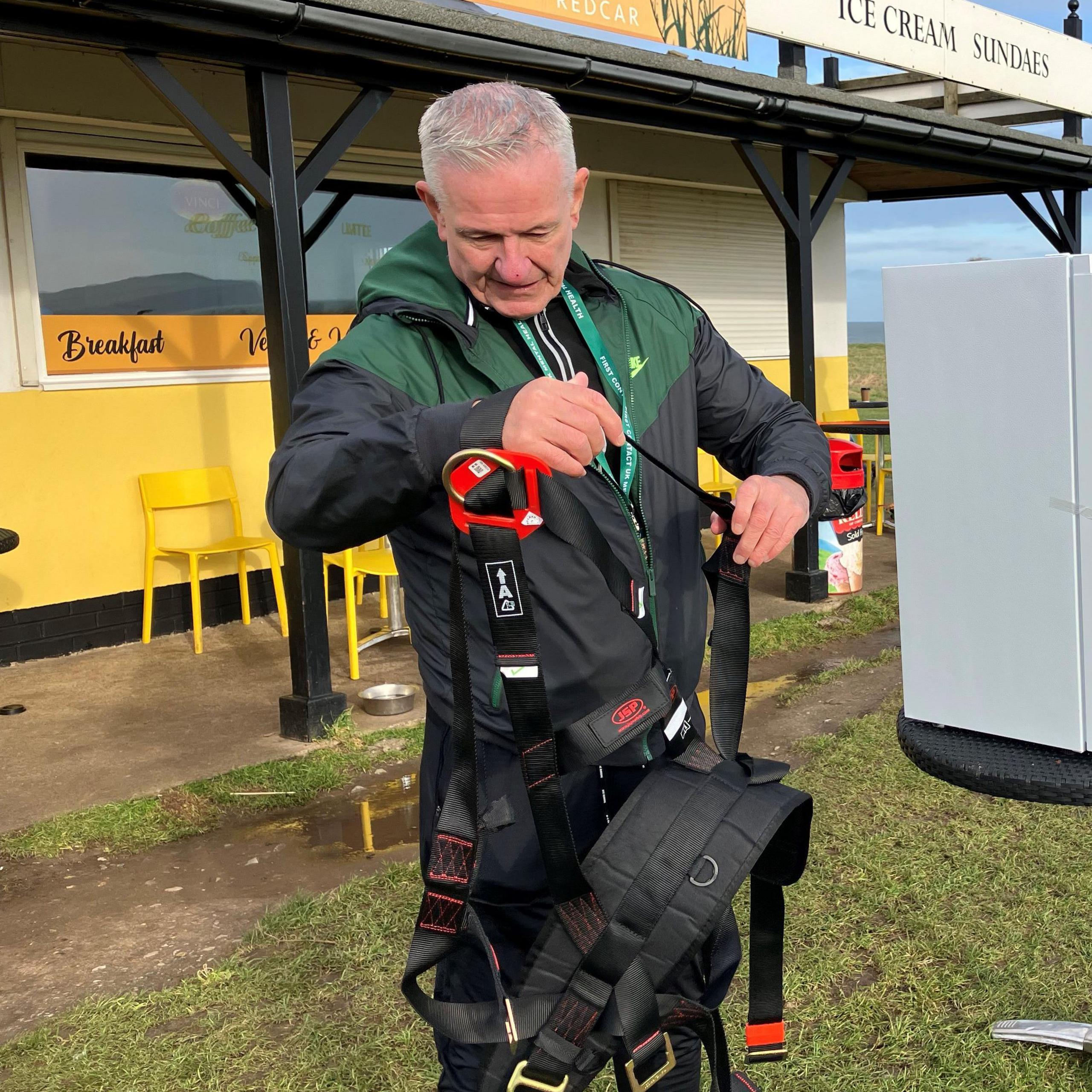 Tom Judge adjusting his straps that he will fasten to the fridge which is to the right of the picture. He is standing on a patch of grass and there is a cafe in the background. 