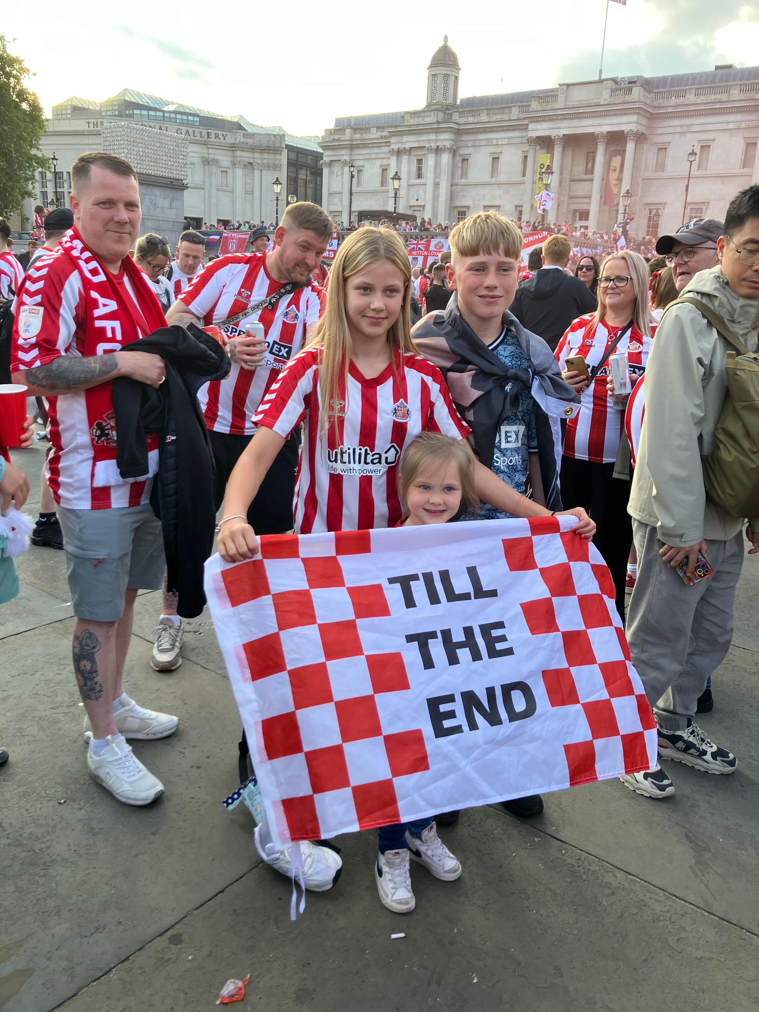 A boy and girl with a little girl and a sign saying Till The End. They are wearing red and white Sunderland shirts and are at Trafalgar Square. Adults are next to them wearing Sunderland shirts.