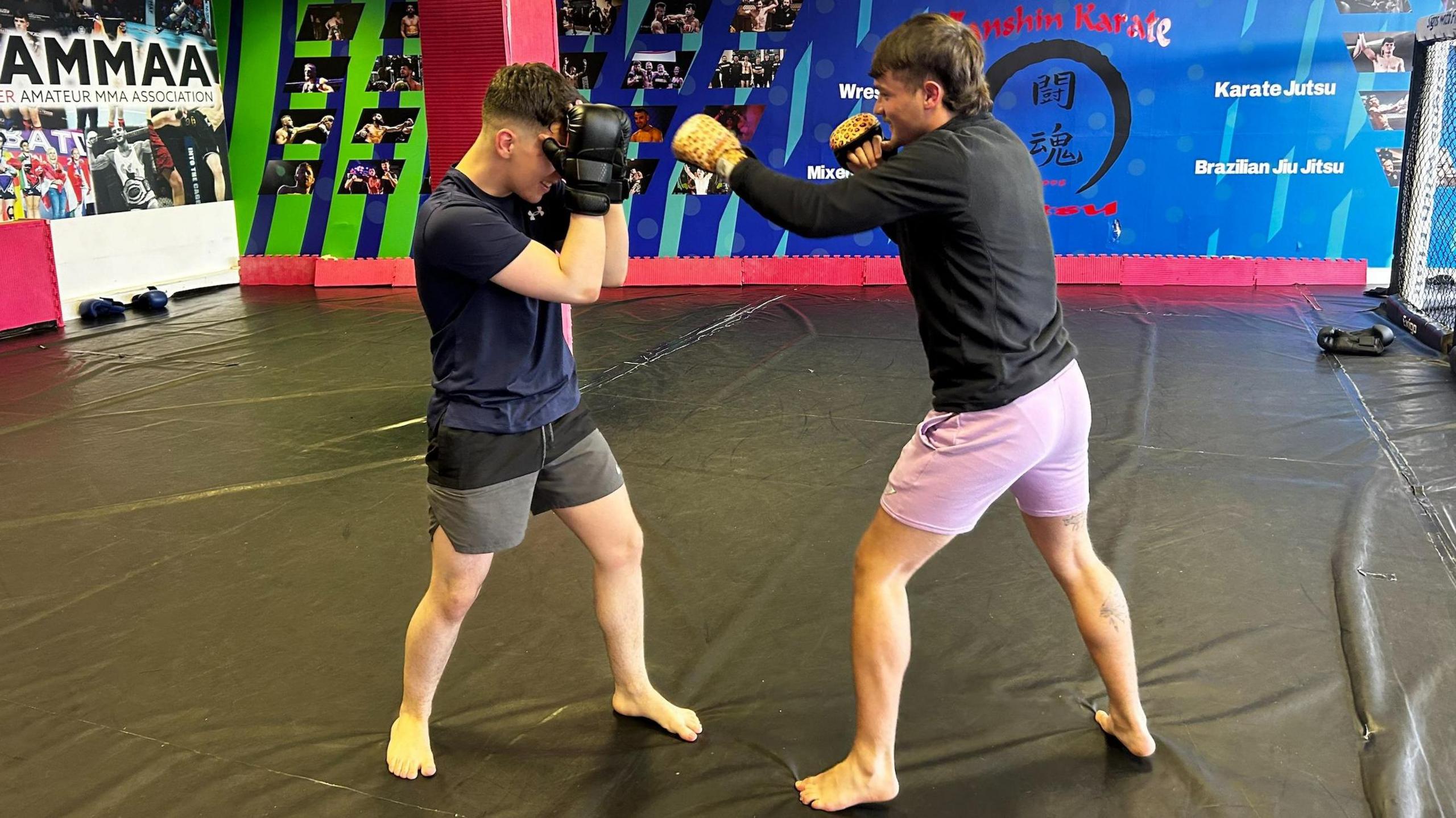 Two young men are sparring. One has lilac shorts and a black top and the other has a navy top and grey shorts.