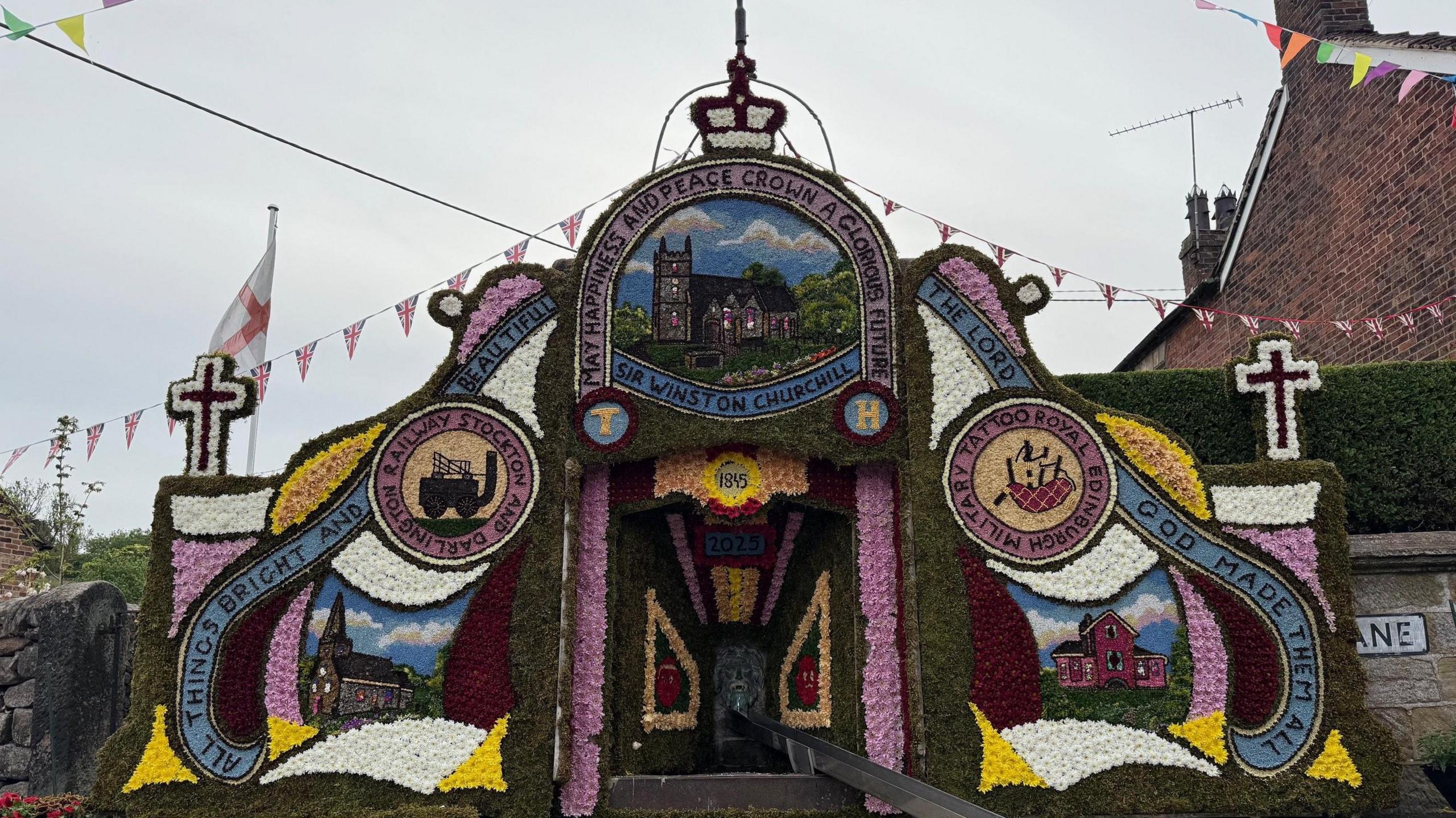 A traditional Derbyshire-style well dressing, featuring an array of colourful designs surrounding a stone well. There is bunting around the well and houses in the background.