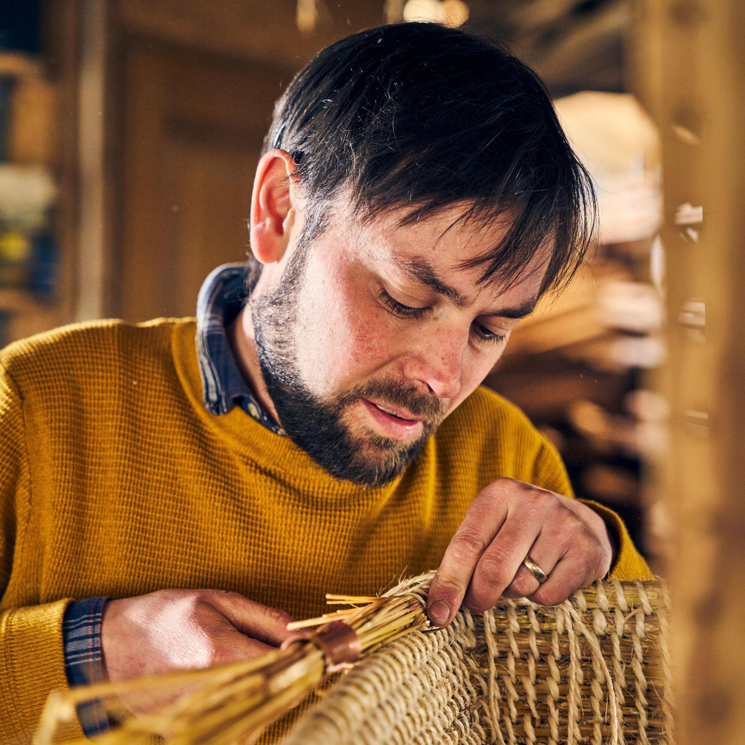 A close-up of bearded Orkney chair maker Kevin Gauld, leaning over a chair in construction, working with his hands to weave the structure. He is a study in concentration, with a mustard-coloured jumper over a checked shirt.