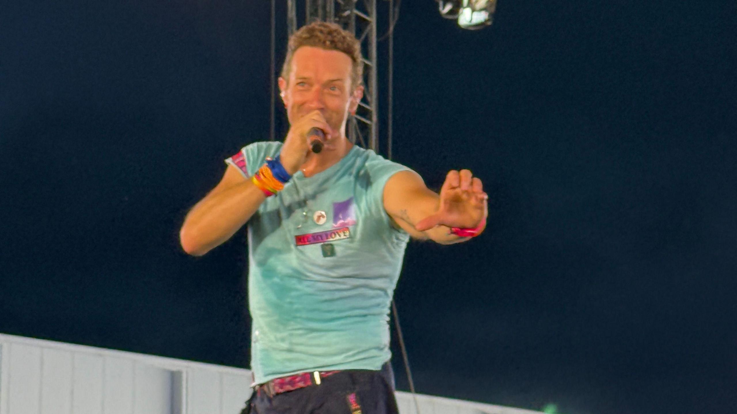 Coldplay lead singer Chris Martin holds his left arm out while holding a microphone in his right hand during a concert. He has a turquoise t-shirt on which has some badges on it. He is wearing something with a pink strap on his left wrist and multicoloured bands on his right wrist.