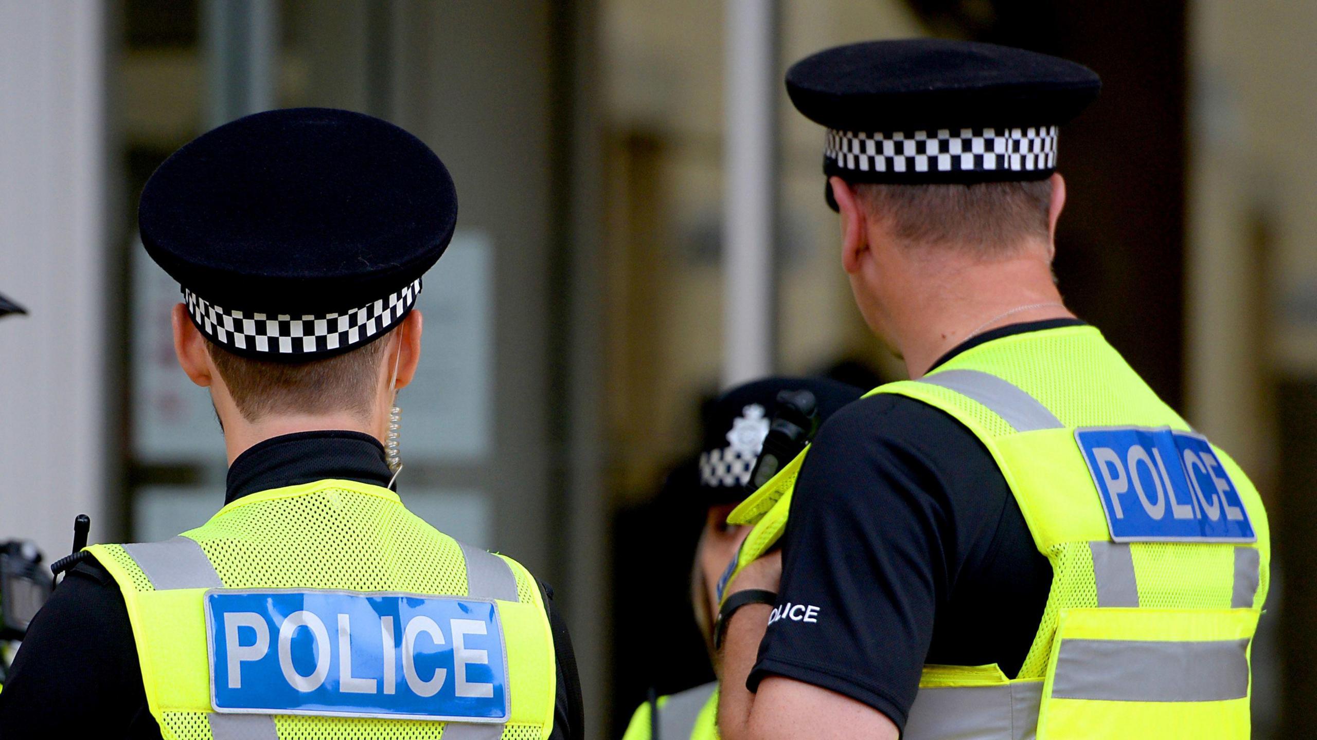 Two police officers with their backs to the camera, wearing police hats and hi-vis jackets that say 'police' on a blue badge