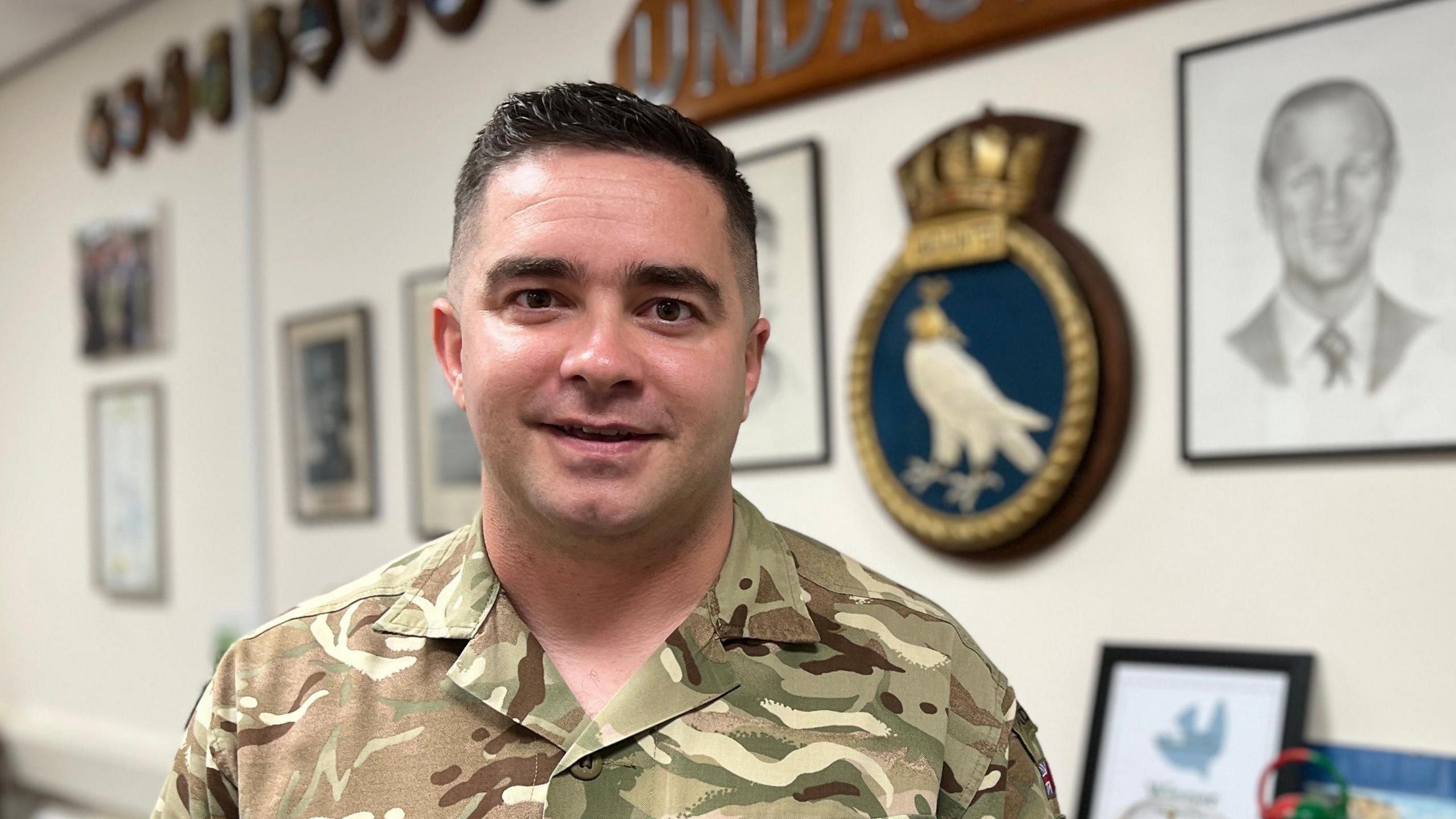 Craig is in camouflage military uniform stands in an office-like room with framed photographs, plaques, and a large wooden sign reading 'UNDAUNTED' featuring a bird crest. A table with various objects, possibly models or decorations, is visible in the foreground.