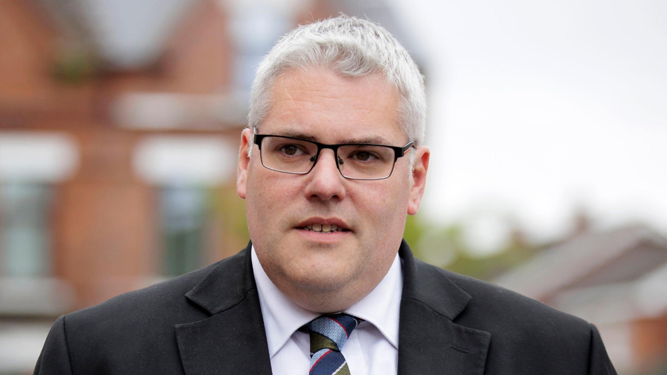 Gavin Robinson pictured outside wearing a grey suit, green tie and spectacles. 