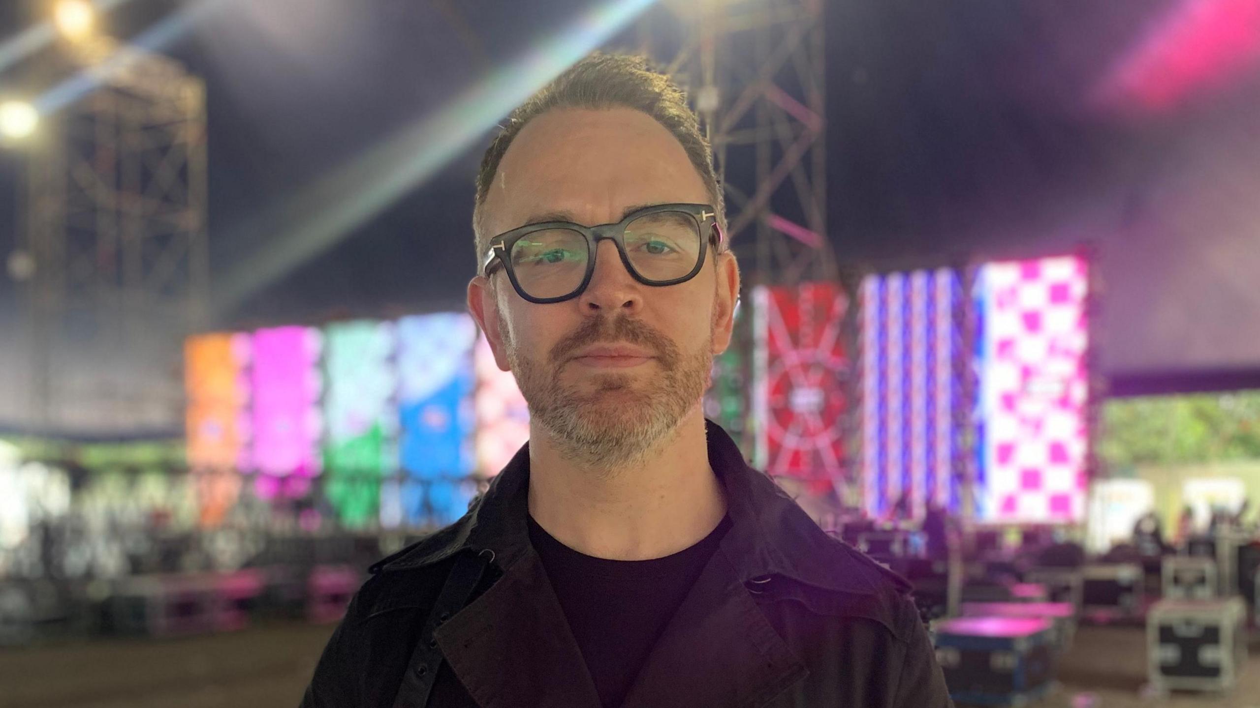 Joe is standing in the Shine tent at the Emerge venue. He is looking at the camera. He is wearing a black top and a black jacket and glasses. Behind him, there is a screen showing different colours.