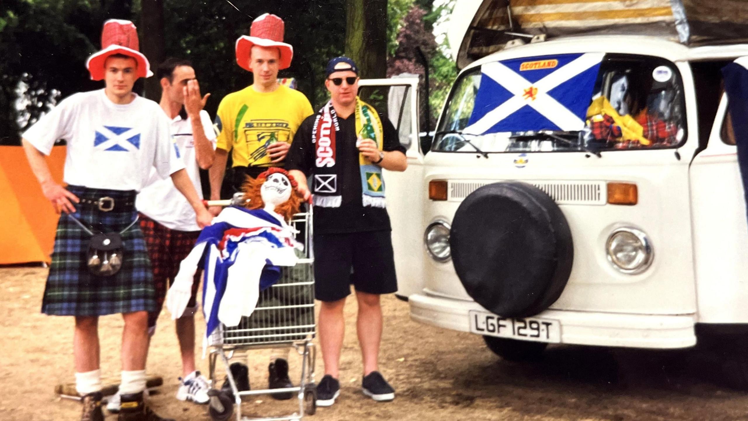 A young Ian Stephen in a kilt, a Scotland saltire T-shirt and a silly red hat, stands beside three similarly-dressed friends in 1998, with a shopping trolley containing a skeleton in a "see you Jimmy" hat, beside their 1979 VW campervan