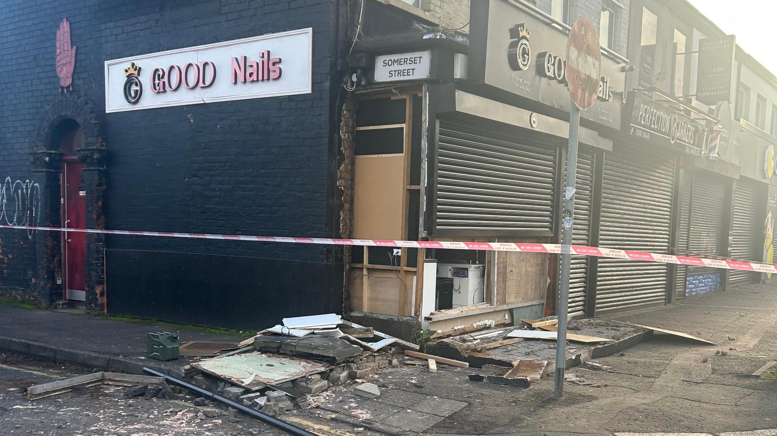 Damage at Good Nails which is on the corner of a street. The side of the shopfront is completely caved in and there is debris along the footpath. A red police cordon is all along the footpath. The side of the building is painted black with a red door and a red hand of Ulster on the wall. A sign for Somerset Street is on the side of the building.