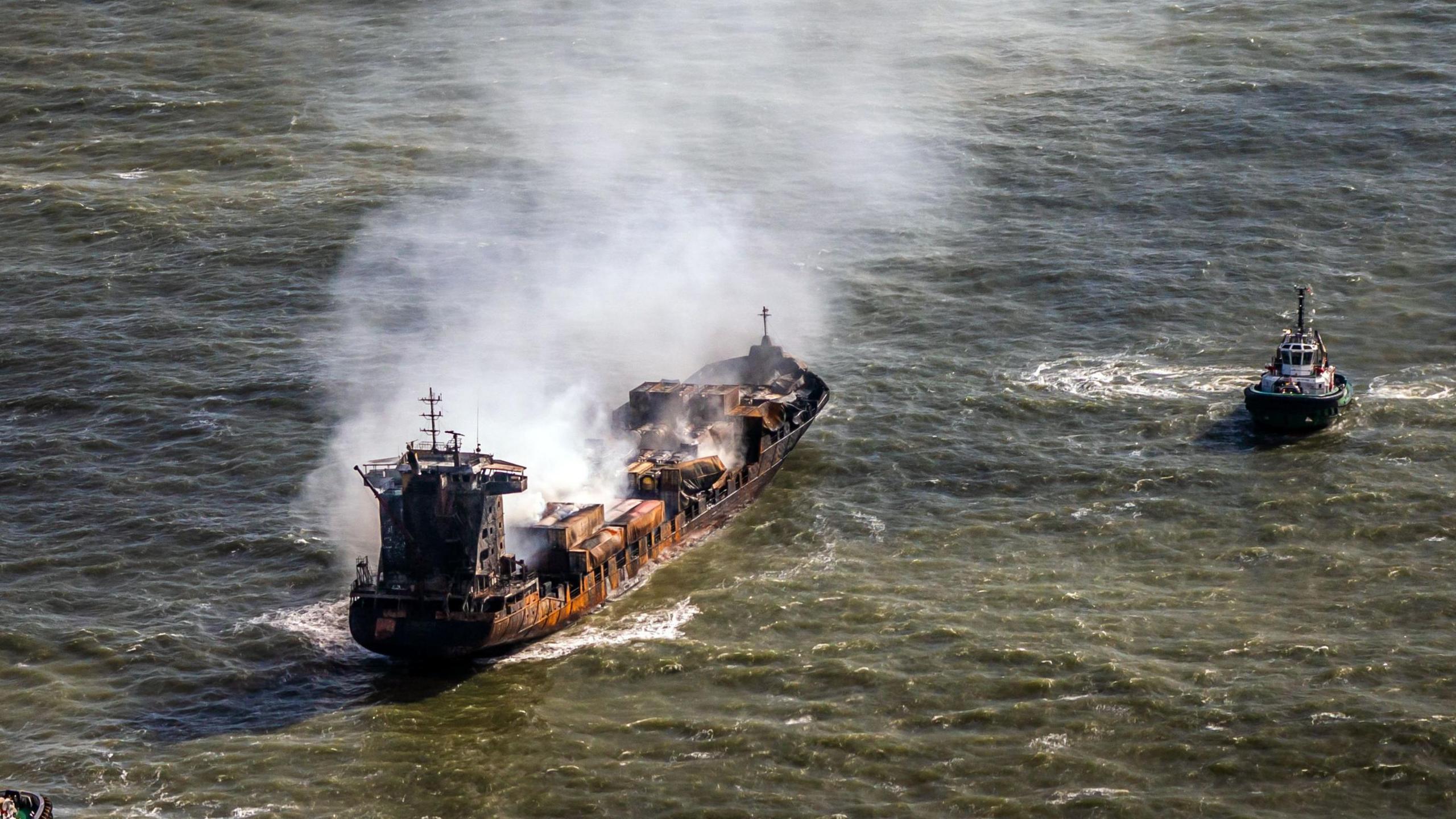 Tug boats shadow the Solong container ship as it drifts in the Humber Estuary, off the coast of East Yorkshire following a collision with the MV Stena Immaculate oil tanker. Smoke can be seen billowing from the ship which has char and burn marks on it. 