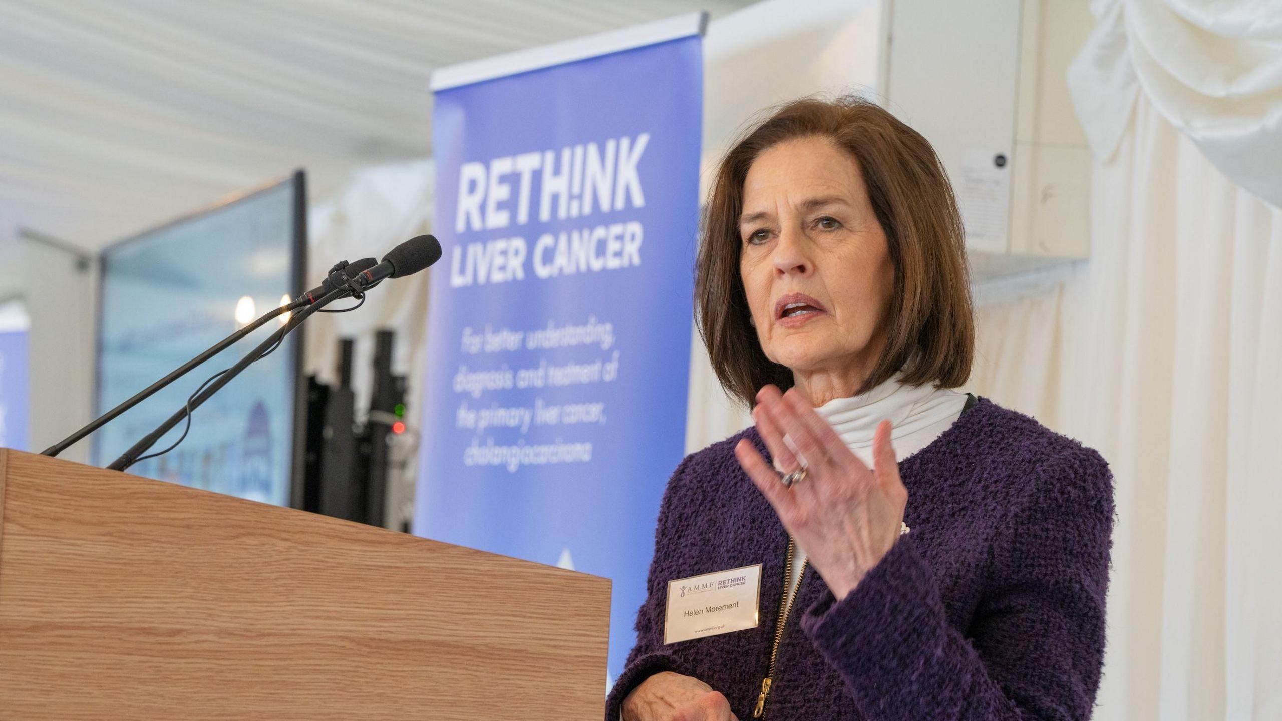 AMMF chief executive, Helen Morement gives a speech in front of a a purple banner that says "rethink liver cancer." She has brown bobbed hair and wears a white roll neck jumper under a purple zip-up jacket.